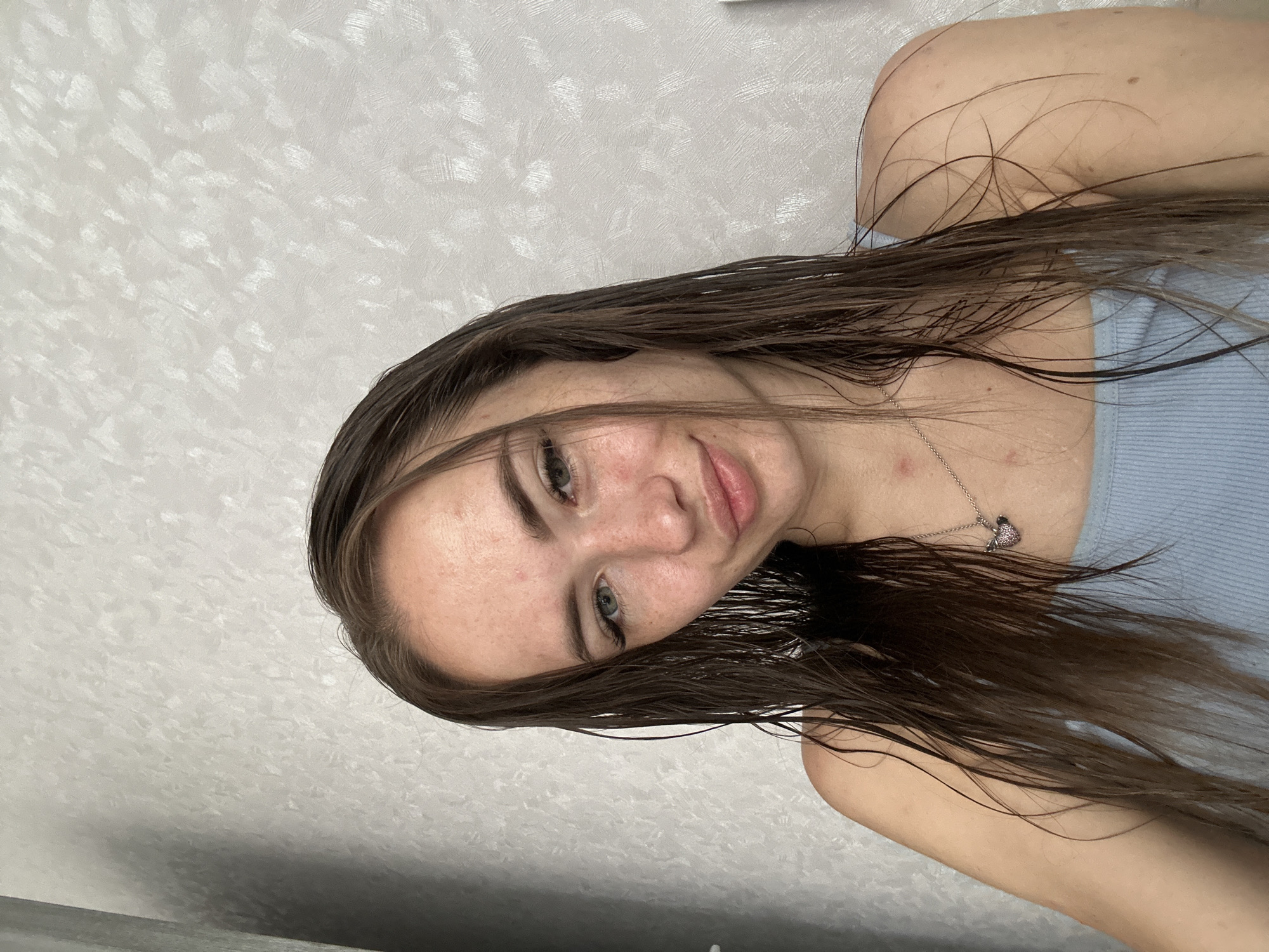 A young woman with wet, brown hair and freckles poses for a selfie against a textured, light-colored wall. She is wearing a light blue tank top and a silver necklace with a heart pendant. Her skin has some redness, particularly on her cheeks. The lighting is soft, casting a shadow on the left side of the frame.