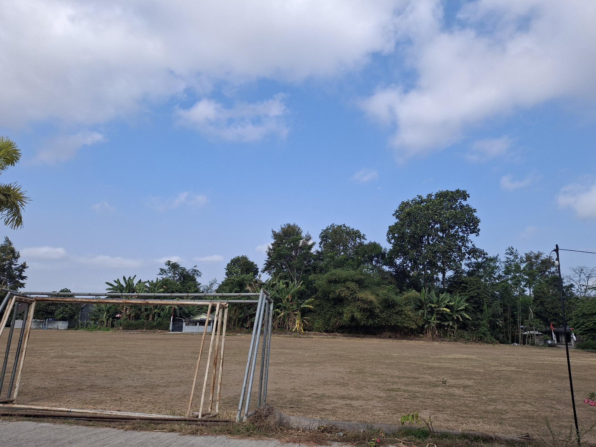 This image depicts an outdoor scene in Labruk Kidul, Indonesia, featuring an empty dirt field with a dilapidated metal soccer goal in the foreground. The field appears dry and somewhat barren. Behind the field, a dense line of trees and vegetation forms a backdrop, with some glimpses of buildings and structures peeking through the foliage. The sky above is a clear blue, dotted with scattered white clouds. The overall impression is one of a rural or semi-rural setting, possibly a school or community ground. There are no people visible in the image.