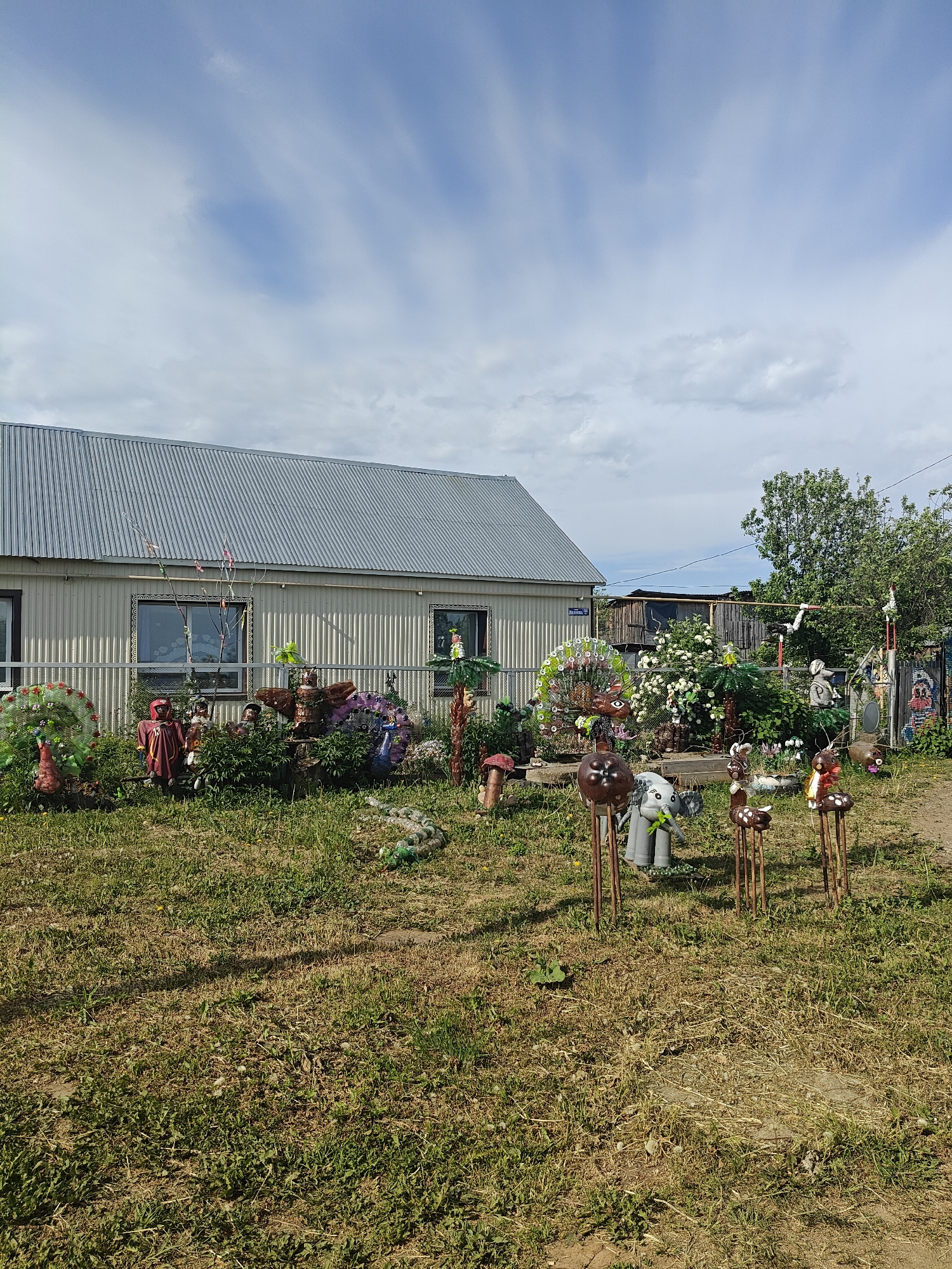 The image depicts a house in Фоки, Russia, surrounded by a yard decorated with numerous whimsical, folk-art sculptures and decorations. The house is a simple, one-story structure with a metal roof and light-colored siding. Two windows are visible on the side facing the camera, framed with decorative lace curtains. A chain-link fence runs along the front of the house.
The yard is covered in grass that appears dry in places. A variety of decorations are scattered throughout, including:
* Animal sculptures (e.g., peacocks, deer, elephants) made from recycled materials such as plastic bottles and tires.
* Figures that appear to be scarecrows or dolls dressed in colorful clothing.
* Mushroom-shaped sculptures.
* Palm tree-like decorations.
* A sculpture that resembles an alligator made from recycled bottles.
* Other unidentified decorative items.
The background includes glimpses of another building partially obscured by trees and bushes. The sky is overcast with a mix of clouds. The scene suggests a rural or semi-rural setting. The decorations indicate a personalized and artistic yard.