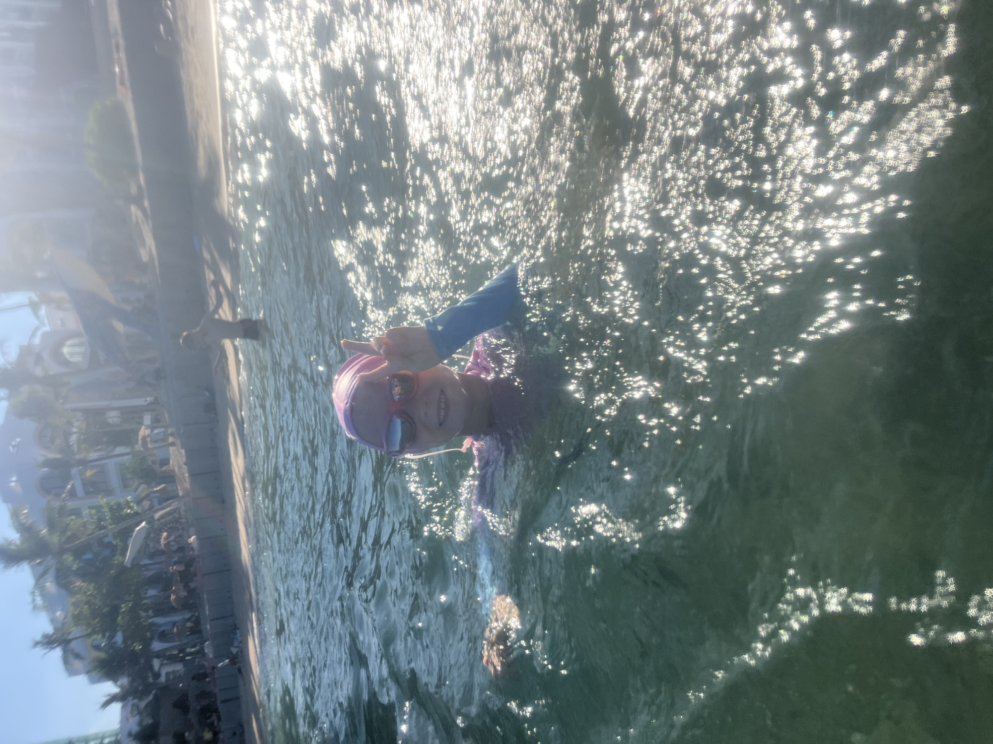 The media file shows a young person swimming in the ocean, viewed from above and at an angle. The person is wearing a purple swimsuit, goggles, and a pink swim cap. They are smiling and making a peace sign with their fingers. The water is choppy and reflects the bright sunlight, creating a sparkling effect. In the background, there is a blurred view of a shoreline with buildings and trees. The image appears to have been taken on a sunny day, likely in the morning or afternoon, given the bright sunlight. The location is identified as Bac Nha Trang Ward, Vietnam. The overall mood of the image is cheerful and playful. There is no visible text in the image.