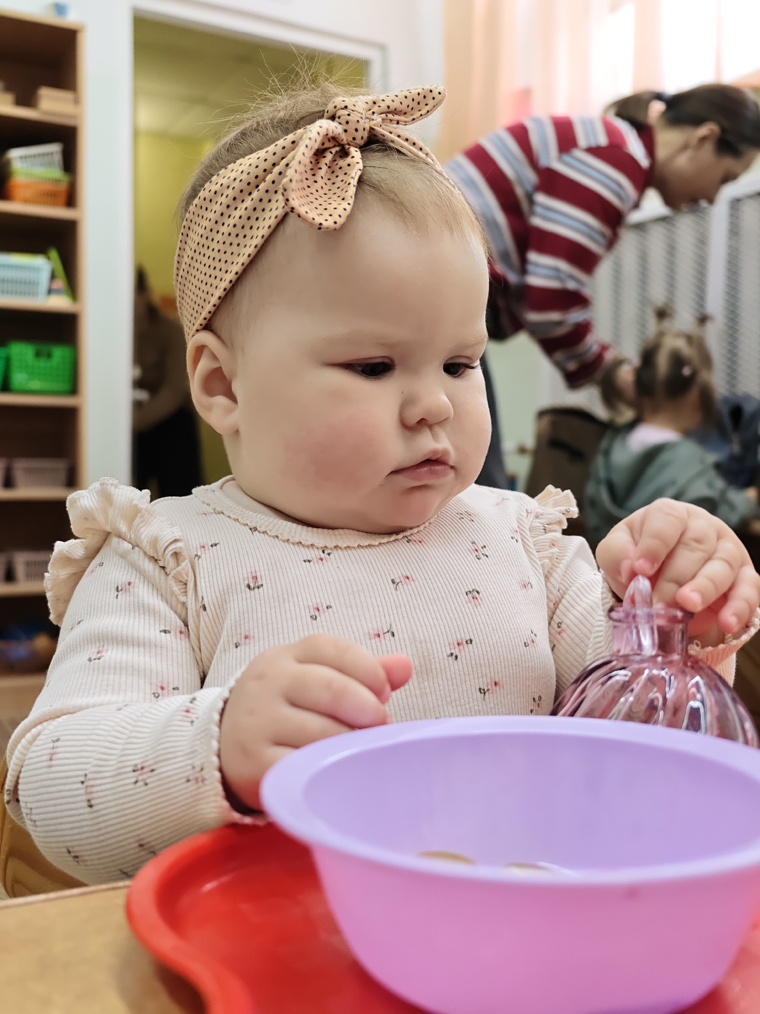 The image depicts a young child, likely a baby or toddler, seated at a table and engaged in an activity within an indoor setting, possibly a daycare or early learning center in Chaikovsky, Russia. The child has light hair and wears a beige headband with black polka dots and a bow. Her expression is one of focus and contemplation as she looks downwards. She is dressed in a light-colored, long-sleeved top featuring subtle floral patterns and ruffles on the shoulders.
In the foreground, a red tray holds a purple bowl containing a few small, round, light-colored objects, possibly tokens or coins. The child holds a small, fluted, light-purple glass bottle or container with a stopper in her right hand, seemingly interacting with the items or the bottle itself as part of a fine motor skill activity.
The background is softly blurred, revealing parts of the environment. To the left, a wooden shelving unit is visible, filled with organized items in colored baskets (green and orange), indicating a structured play or learning area. In the mid-ground, a blurred figure of an adult, dressed in a red and white striped shirt, is seen bending over. Further back, another child with pigtails is partially visible from behind. The lighting suggests it is daytime. No text is visible in the image.