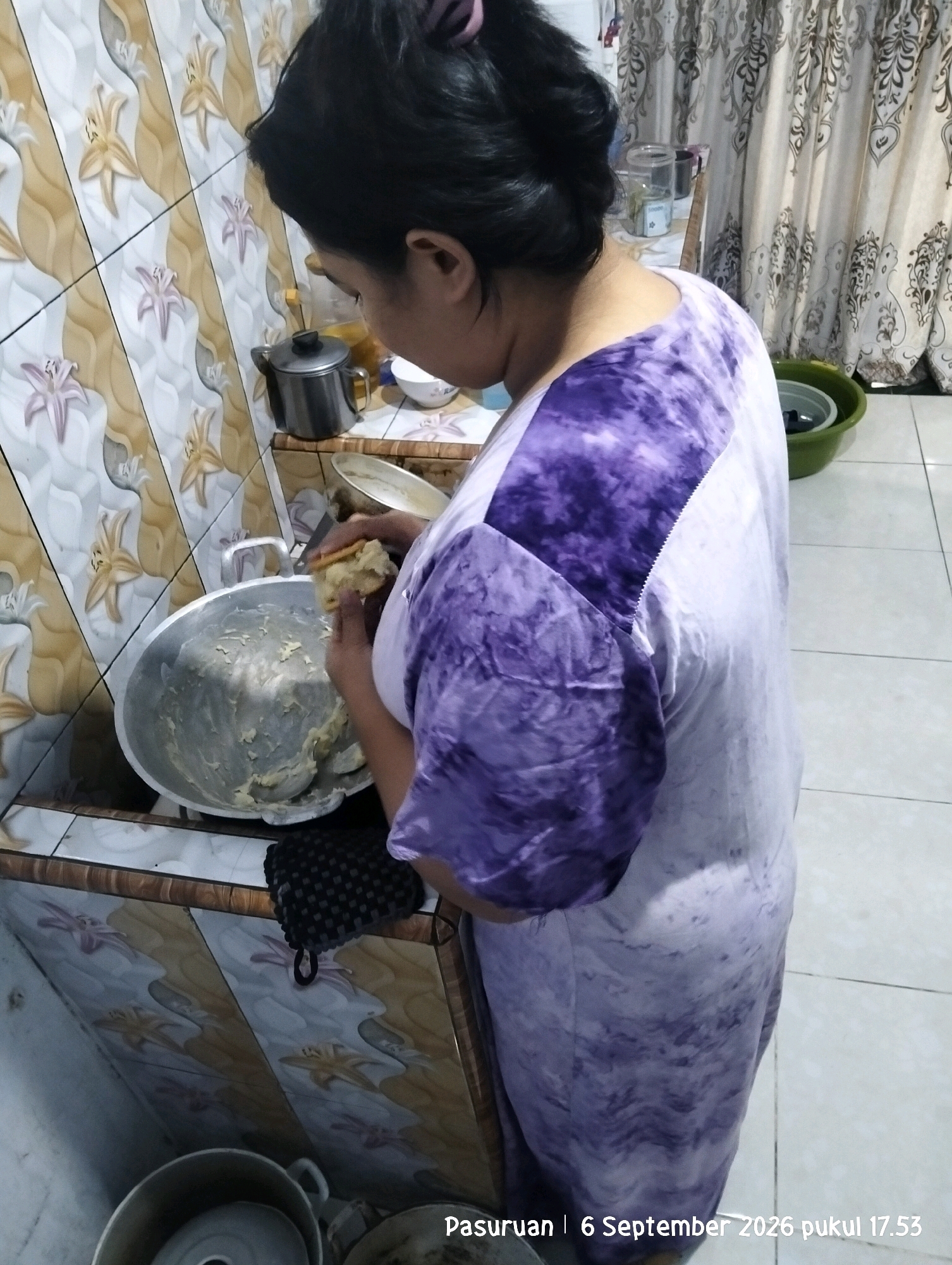A woman with black hair pulled back in a bun is standing in a kitchen, facing away from the camera and looking down into a large, shallow metal bowl. She is wearing a purple and white tie-dye t-shirt. Her right hand is holding a piece of light-colored dough over the bowl, and her left hand appears to be reaching into the bowl. The kitchen has tiled walls with a floral pattern in shades of beige, white, and brown. A silver pot with a black lid is on a shelf to the left of the woman. To the right of the woman, a green plastic basin is visible. The floor is tiled in light gray. A curtain with a damask pattern hangs in the background on the right. A watermark at the bottom right reads "Pasuruan | 6 September 2026 pukul 17.53".