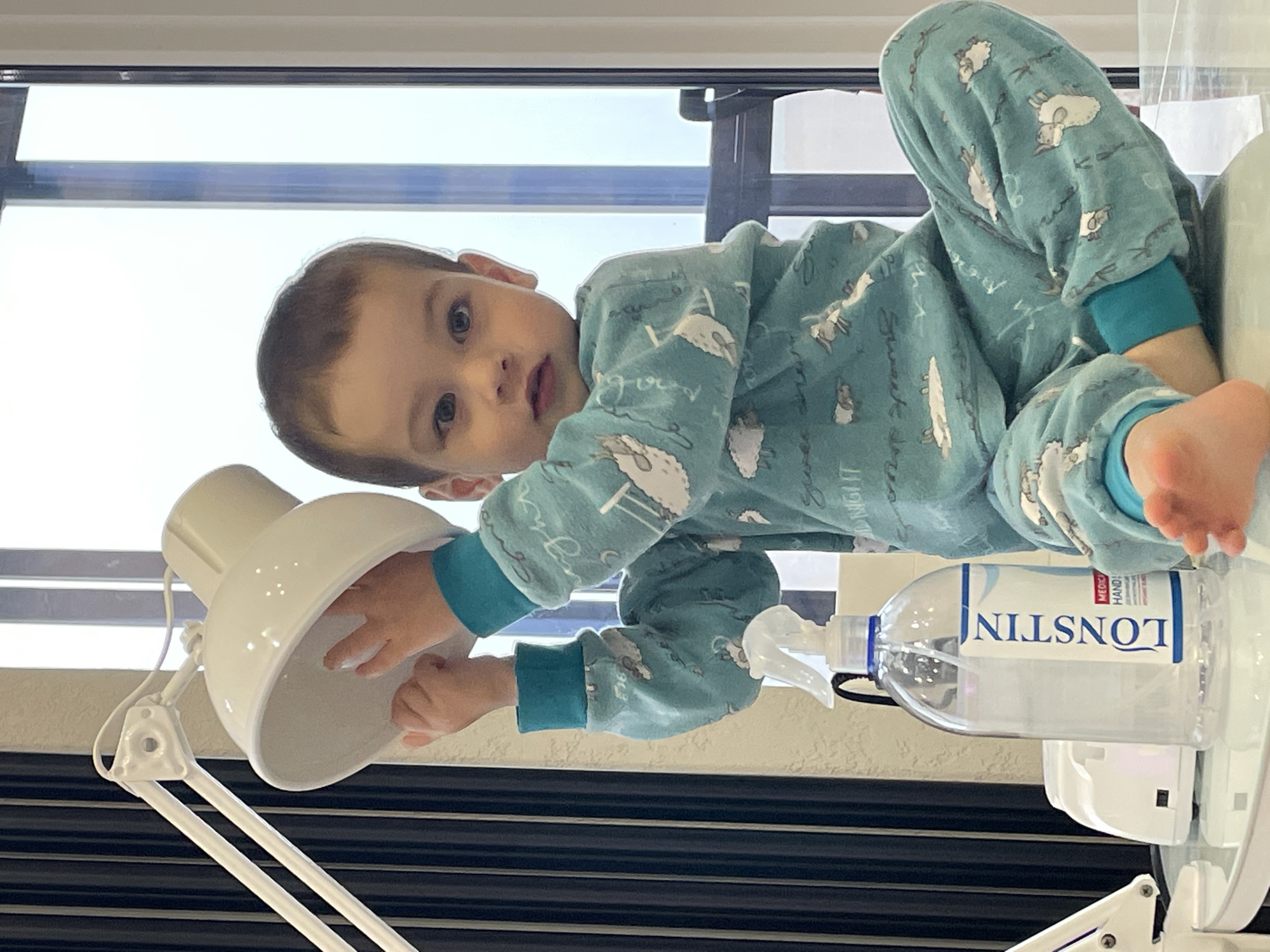 A child wearing patterned pajamas is lying on a surface near a window. The child is reaching towards a white lamp. Next to the lamp is a clear bottle of hand sanitizer labeled "LONSTIN". The location is not identifiable as Izhevsk, Russia from the given image.