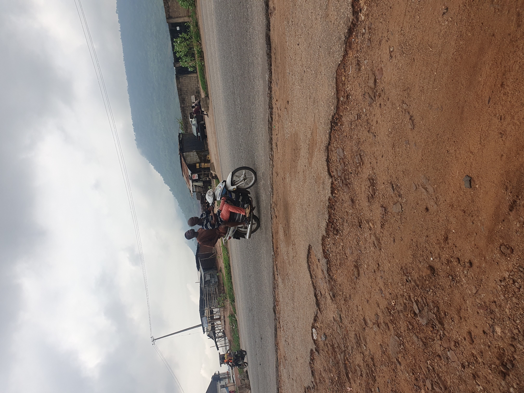 The image shows a street scene in Lokoja, Nigeria. The road is paved but the edges are eroded, exposing red soil. A motorcycle with three people is driving on the road. There are buildings and vegetation along the side of the road. Mountains are visible in the distance. The sky is cloudy.