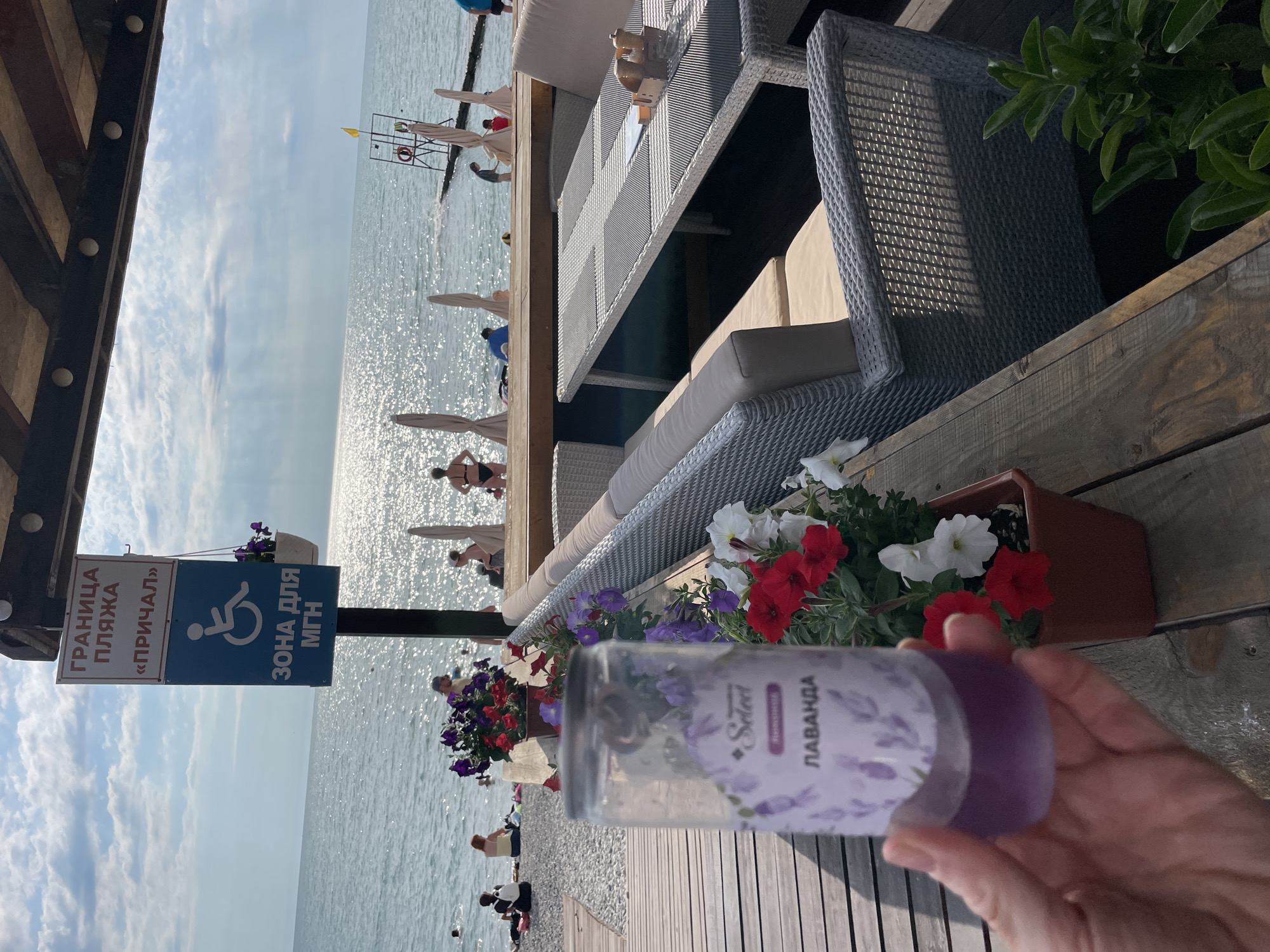 The image captures a sunny day at a beach resort, likely in Sochi, Russia, given the Cyrillic signage. In the foreground, a hand holds a clear plastic container with a purple liquid and a label featuring lavender sprigs and the word "Select" and "Лаванда" (Lavender). The container is placed on a wooden surface, next to a potted plant with red, purple, and white petunias.
Behind the foreground elements, a seated outdoor dining area is visible, featuring a wicker sofa and a table with salt and pepper shakers. The dining area overlooks the Black Sea, where several people are swimming and relaxing on the shore. Beach umbrellas are set up along the coastline.
Further back, a signpost with Cyrillic text reading "ГРАНИЦА ПЛЯЖА «ПРИЧАЛ»" (Beach Boundary "Pier") and a wheelchair accessibility symbol indicates a designated area. The sky is partly cloudy, with sunlight glinting off the water. The overall atmosphere is one of a leisurely summer day at the beach.