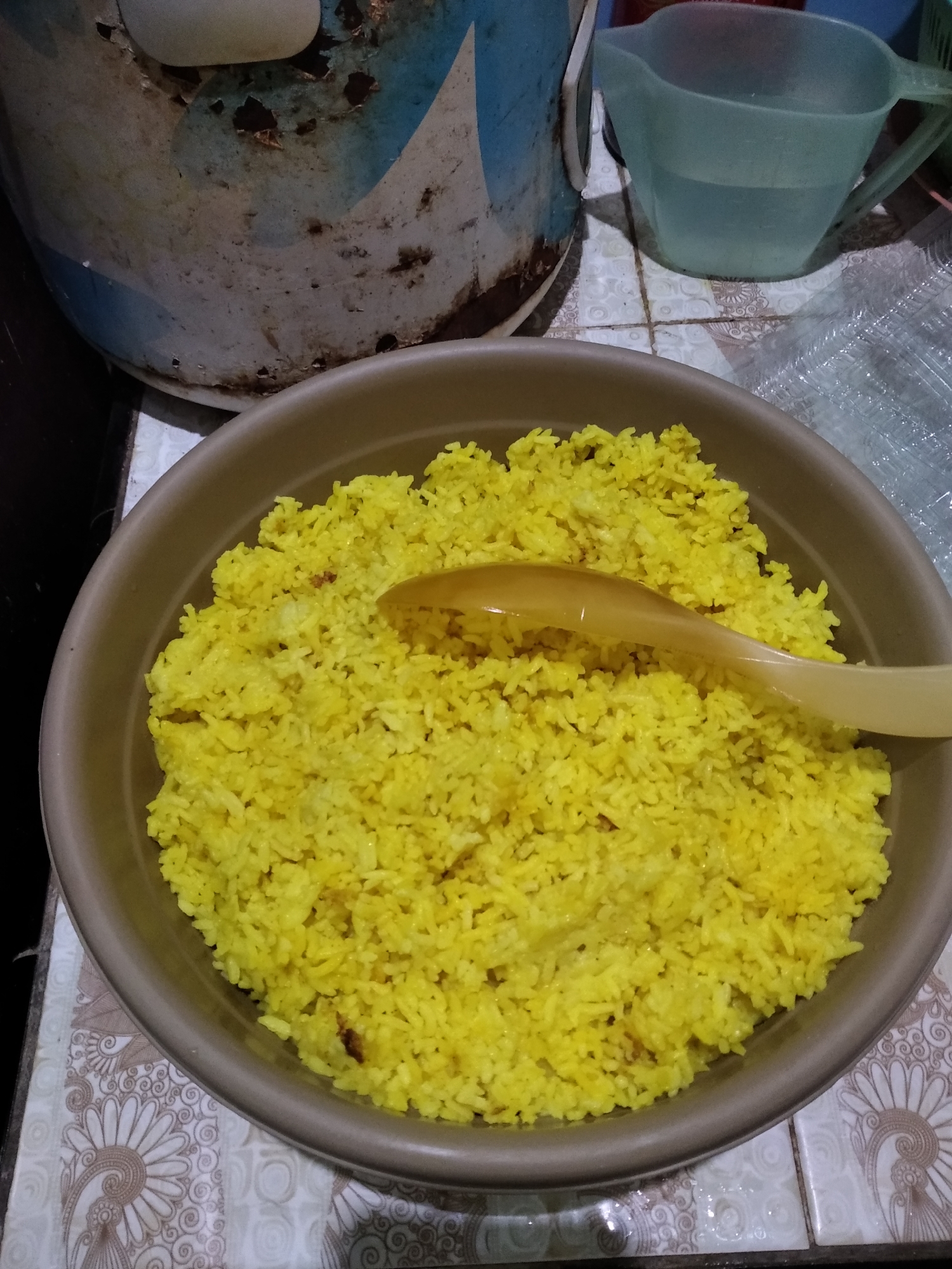 A close-up, top-down shot shows a brown bowl filled with bright yellow rice. A light-colored spoon rests on top of the rice, angled from the upper right towards the center. The rice appears to be cooked and fluffy, with visible individual grains. The bowl is placed on a tiled surface with a decorative floral pattern. In the background, out of focus, is a large, weathered cylindrical object with blue and white markings, possibly a bucket or container. To the right of this object is a clear plastic pitcher filled with a light blue liquid, likely water. The scene is set indoors, likely in a kitchen or dining area. The overall impression is of a simple, homemade meal being served. This scene is located in Rambipuji, Indonesia.