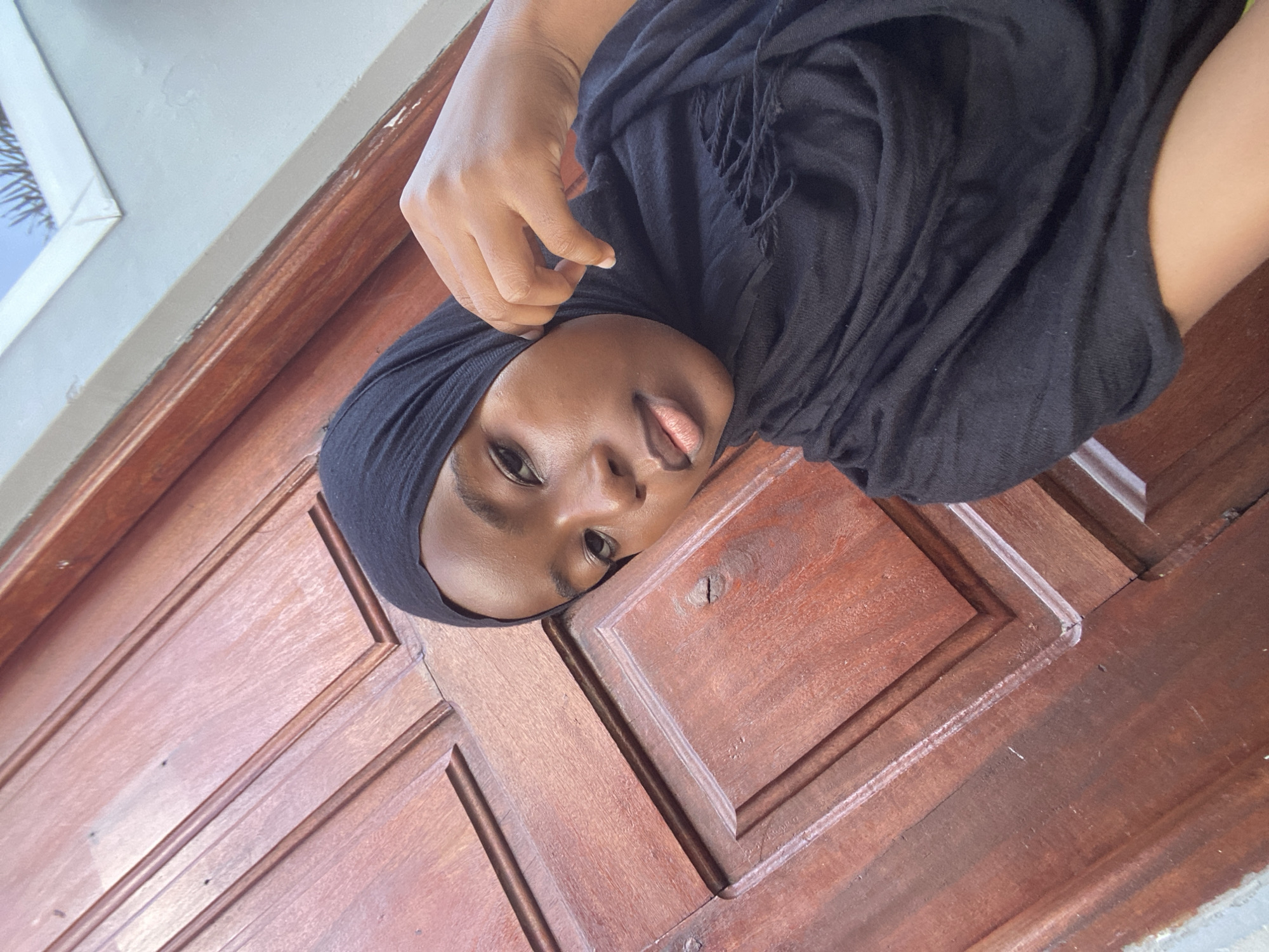 The scene, located in Dar es-Salaam, Tanzania, depicts a young individual with a dark skin tone, captured from the chest up against a textured background. The individual, dressed in a black headscarf and a black garment, faces forward with a neutral expression. Their right hand is positioned with fingers lightly touching the headscarf near the right side of their face. The primary background element is a reddish-brown wooden structure, likely a paneled door or wall, featuring distinct grooves and visible wood grain, with some minor surface imperfections. A portion of a white-painted wall or frame is visible on the far right, adjacent to a light blue area containing faint, spiky outlines. The scene is illuminated by soft, even lighting.