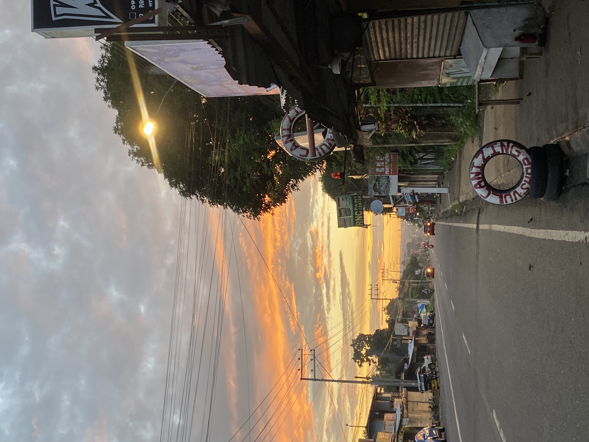 The image captures a roadside scene in Davao City, Philippines, during a vibrant sunset. A two-lane asphalt road stretches into the distance, with vehicles including a blue sedan, a white car, a tricycle, and motorcycles visible. Along the left side of the road, a row of shops with corrugated metal roofs and various signage, including "RNC PAINT CENTER," lines the street. Utility poles with a complex network of wires crisscross the sky.
On the right side of the road, a storefront with a dark, weathered awning is present. A white circular sign with red lettering, reading "CANTING SHOP," is mounted on a pole and also stacked on tires on the sidewalk. A street lamp illuminates the area with a bright, warm glow, and its light flares across the frame. The sky is a spectacle of orange, pink, and gray clouds, indicative of a sunset. Small birds are visible on the road and sidewalk. The overall atmosphere is that of a busy, yet serene, urban environment transitioning into evening.
