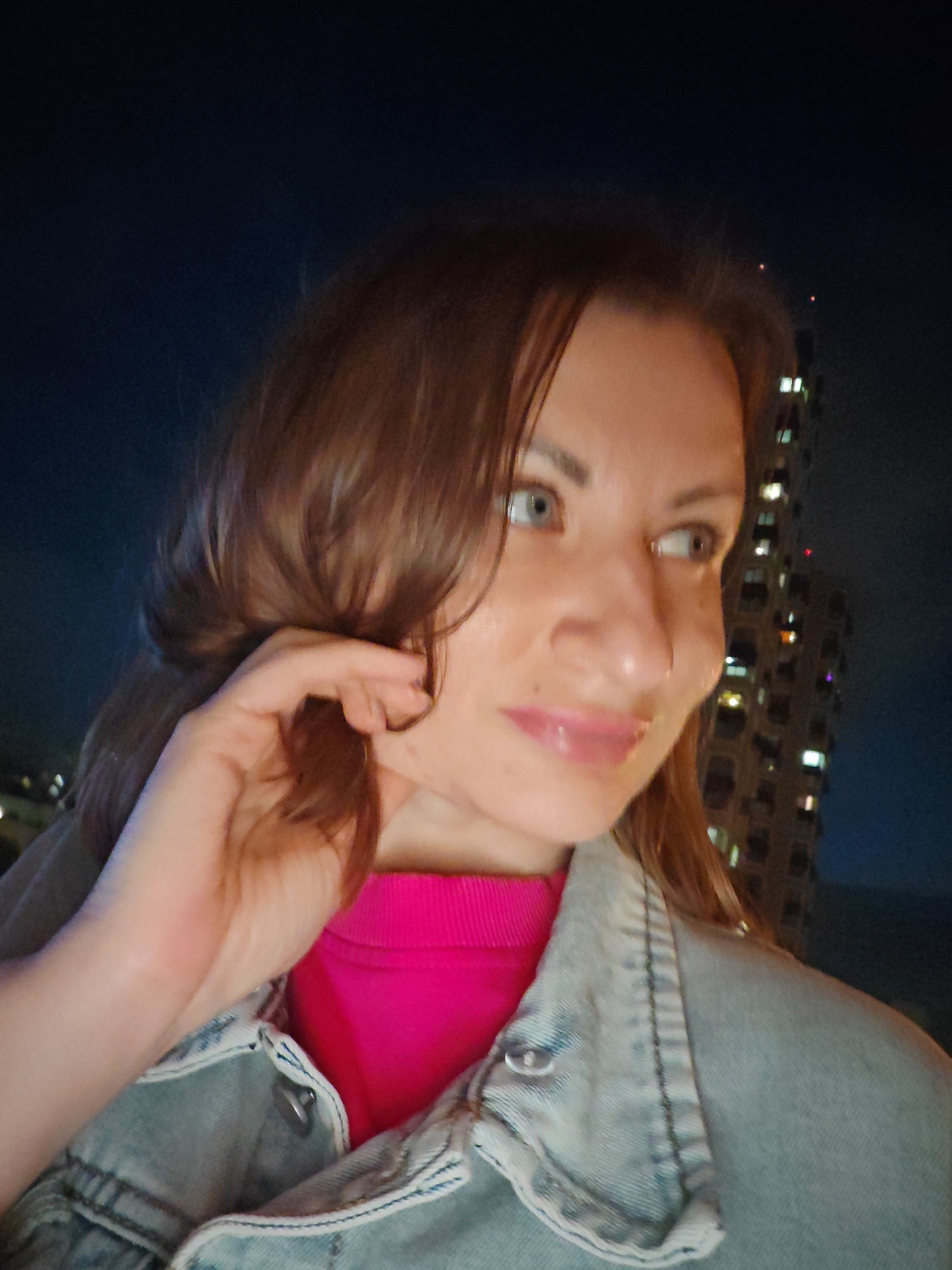 In a nighttime, medium shot taken outdoors, a woman with medium-length brown hair and blue eyes is looking to her right. She is wearing a bright pink shirt and a light blue denim jacket. Her right hand is touching her hair near her face. Behind her, a tall, modern building with many illuminated windows is visible against a dark blue night sky. The setting appears to be an urban environment.