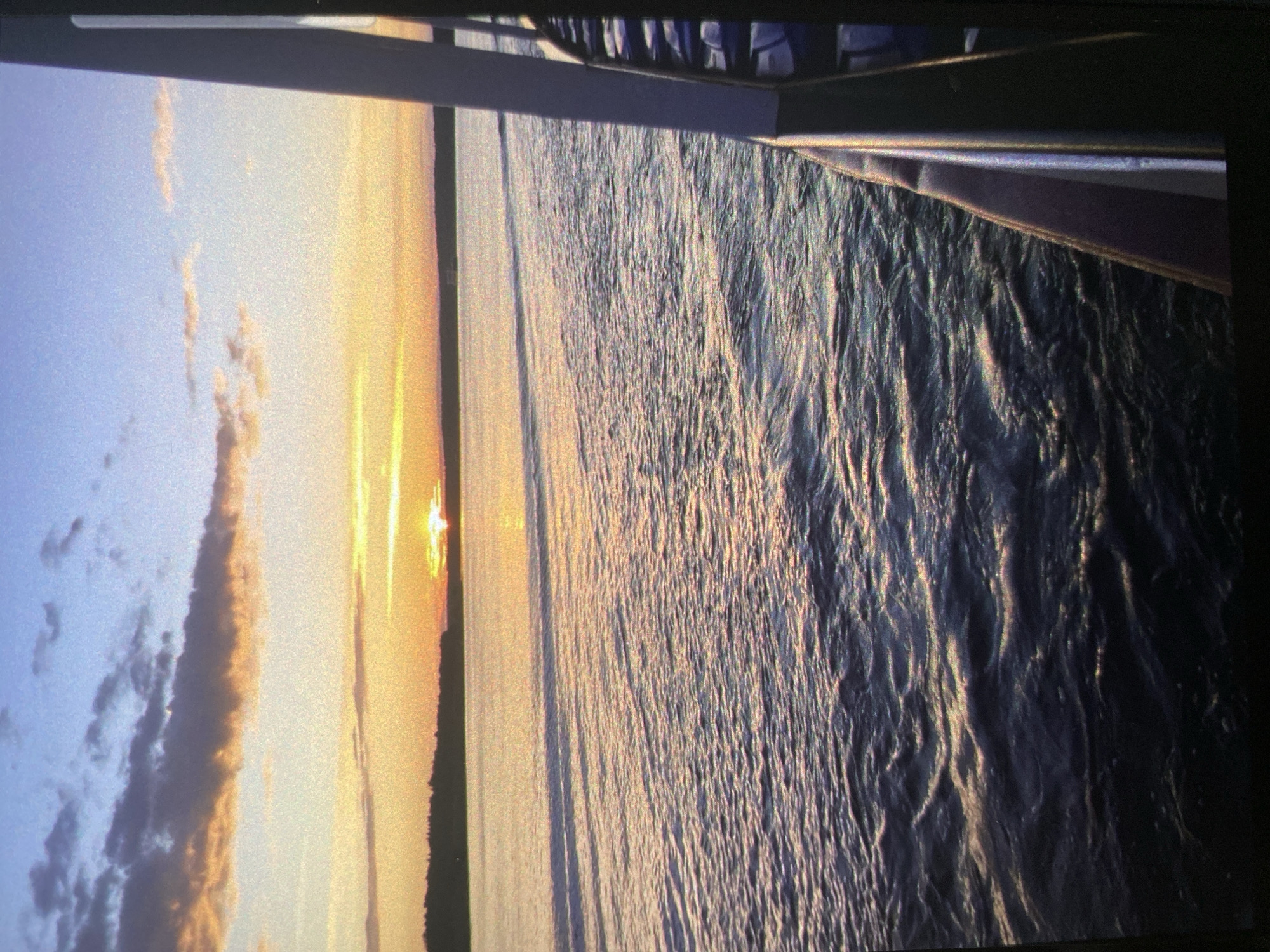 The media file is a landscape photograph, rotated 90 degrees clockwise, depicting a sunset or sunrise over a vast body of water, likely taken from a boat or waterfront structure in Saint Petersburg, Russia.
In the foreground, occupying the top portion of the correctly oriented image, is a dark, horizontal railing or edge, suggesting a vantage point from a vessel or pier. Below this structure, the expansive water surface is visibly rippled and choppy, reflecting the warm light of the low sun. The water transitions from darker, more textured patterns closer to the viewer to a brighter, almost golden sheen further out.
The sun, a brilliant disc of light, is positioned low on the horizon, creating a luminous path across the water. A dark, silhouetted stretch of distant land or coastline is visible directly beneath the sun. Above the horizon, the sky displays a gradient of colors, transitioning from a soft blue on the left to intense yellows, oranges, and pinks around the sun. Several elongated clouds, some appearing wispy and others denser, are scattered across the sky.
The time of day is clearly either sunrise or sunset, characterized by the dramatic warm lighting and the sun's position. The weather appears clear and calm, with gentle waves on the water. No people, identifiable objects other than the railing, or visible text are present in the image. The scene evokes a sense of peaceful natural beauty.