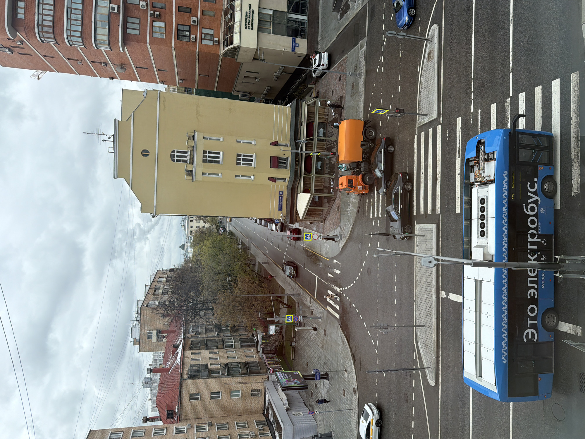 An aerial view from a building captures a bustling street scene in Moscow under an overcast sky. The asphalt appears damp, suggesting recent rain. In the foreground, a blue electric bus, prominently featuring "Это электробус" (This is an electrobus) and "490006" on its side, is positioned near a zebra crossing. The bus also has a distinctive wavy pattern on its upper side.
Various vehicles populate the wet road, including two orange utility trucks, a blue car, a white taxi with yellow stripes, and several dark-colored cars. Pedestrians can be seen walking on the sidewalks and near crosswalks.
The street is lined with multi-story buildings. To the left, a tall red brick building with curved balconies dominates the upper part of the frame. Below it, a prominent yellow building features a ground-floor establishment with the sign "MICHEL" and ornate windows. Another building displays partial Russian text: "ОТ УЛИЦЫ АМАЛИИ КАЛИНЫХ..." (From Amalia Kalinina Street...). On the right side of the street, more brick buildings are visible, interspersed with bare trees, hinting at autumn or early spring. Overhead power lines crisscross the cloudy sky. The scene depicts typical urban activity with traffic lights and road signs visible throughout.