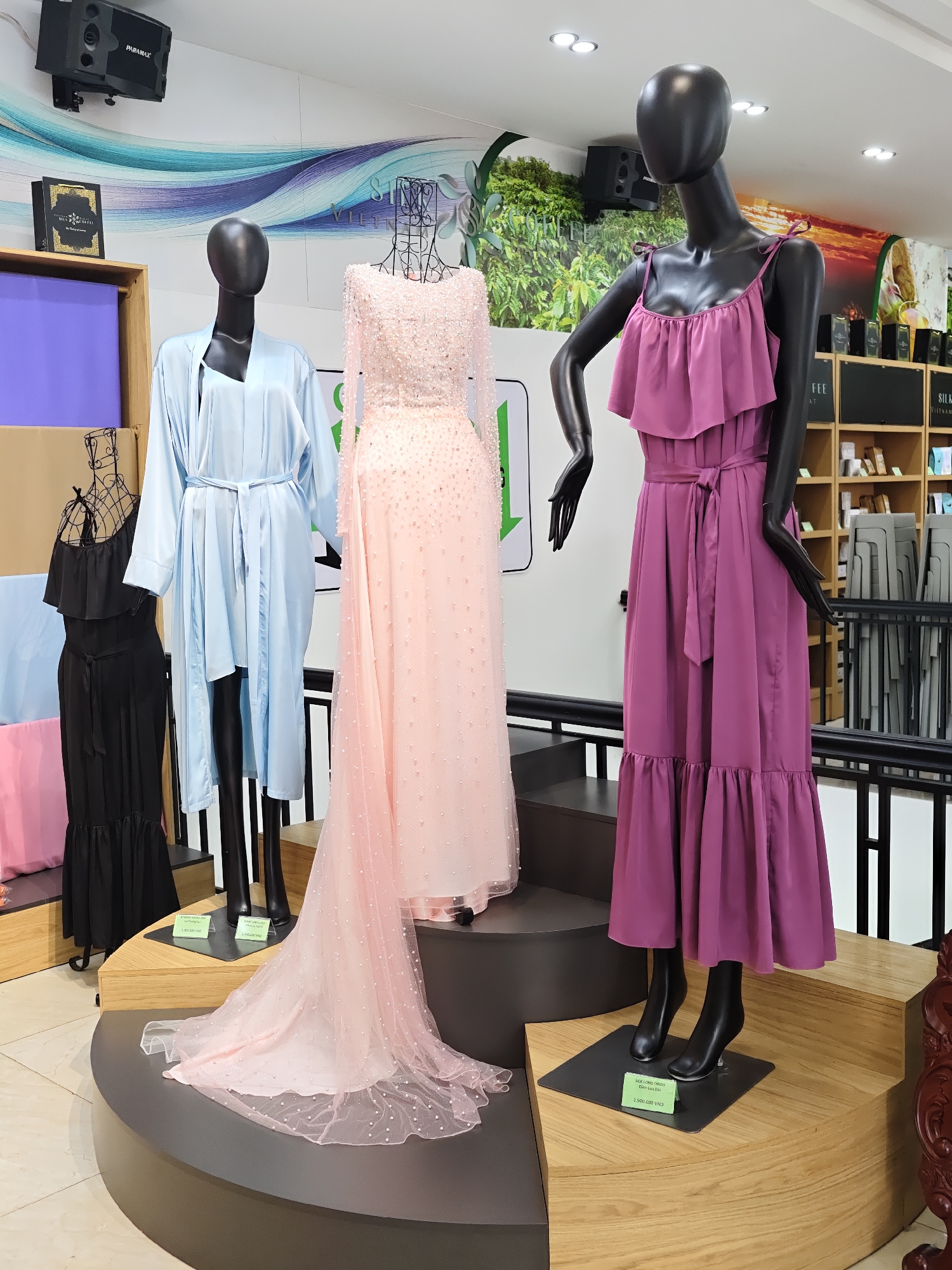 The image captures a retail display in Lam Vien - Da Lat, Vietnam, featuring several mannequins adorned with women's clothing. In the foreground, a black mannequin is dressed in a long, ruffled, magenta dress with a tie waist and thin straps. To its left, another mannequin wears a pale blue, satin robe with a matching belt. A black, tiered, ruffle-bottom dress is displayed on a wire mannequin to the left of the blue robe. A third mannequin, positioned centrally and slightly behind, is adorned in a long, pale pink gown embellished with pearls and beads, featuring a flowing train that extends onto the display platform. The mannequins are arranged on a multi-level, circular wooden display platform. In the background, shelves are stocked with various products, suggesting a boutique or department store setting. A large, abstract mural with blue and white wavy lines adorns the wall behind the display. Additional elements include a black speaker mounted on the wall and lighting fixtures on the ceiling.