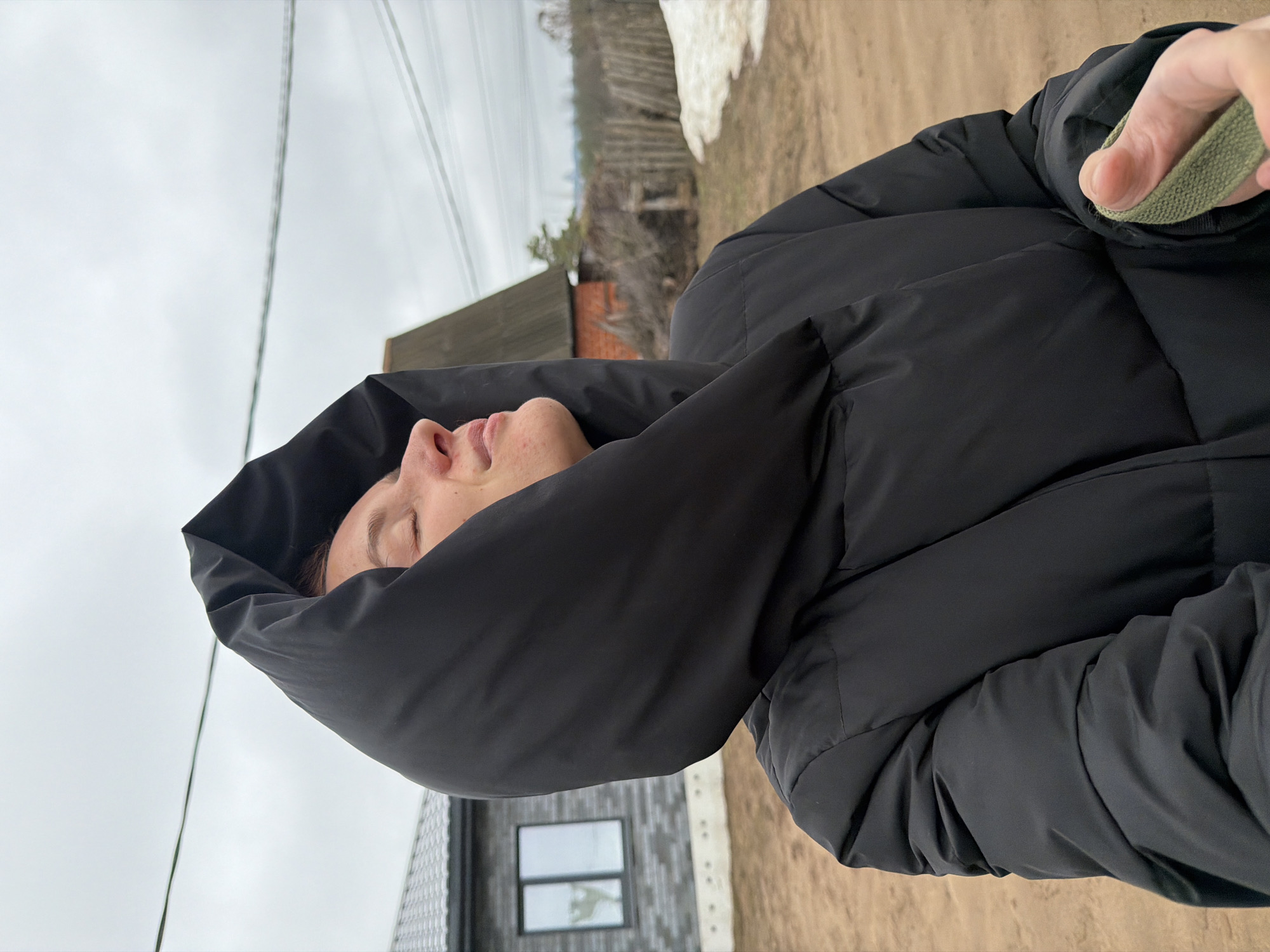 A woman is shown in a medium, angled close-up shot, wearing a black, hooded puffer jacket. She has her eyes closed and her head tilted back. A hand holding a green strap is visible near her right shoulder. The background reveals a cloudy sky, electrical wires, a snow-covered landscape, and buildings, including a brick structure and a dark gray building with a window. The ground appears to be dirt or gravel. The location is potentially the city of Chaikovsky, Russia.