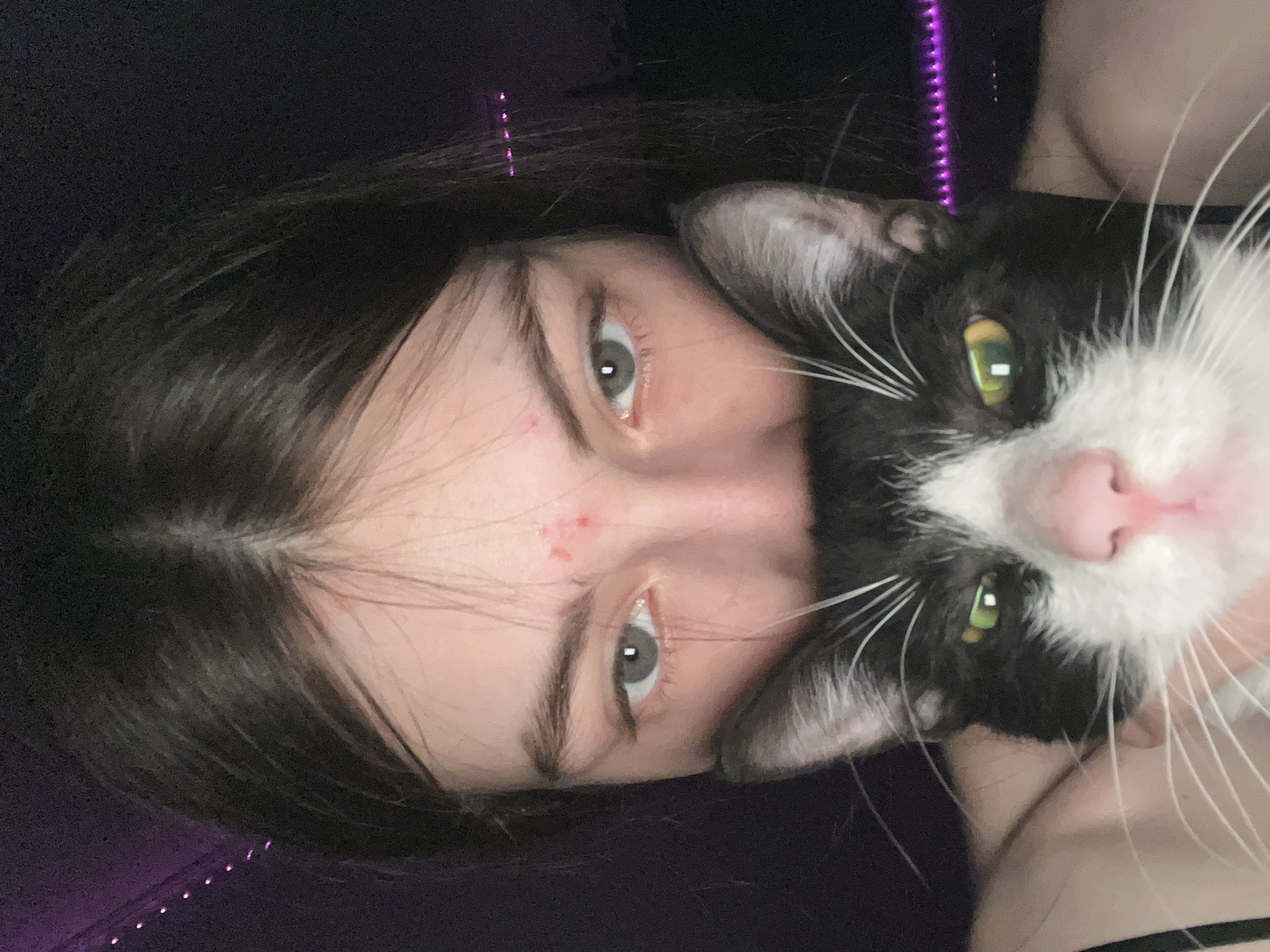 A young woman with dark hair and grey eyes is holding a black and white cat with green eyes close to her face. The background is dark with faint purple lighting. The photo is taken at an angle, making the subjects appear sideways. There is no indication of the location being Saint Petersburg, Russia.