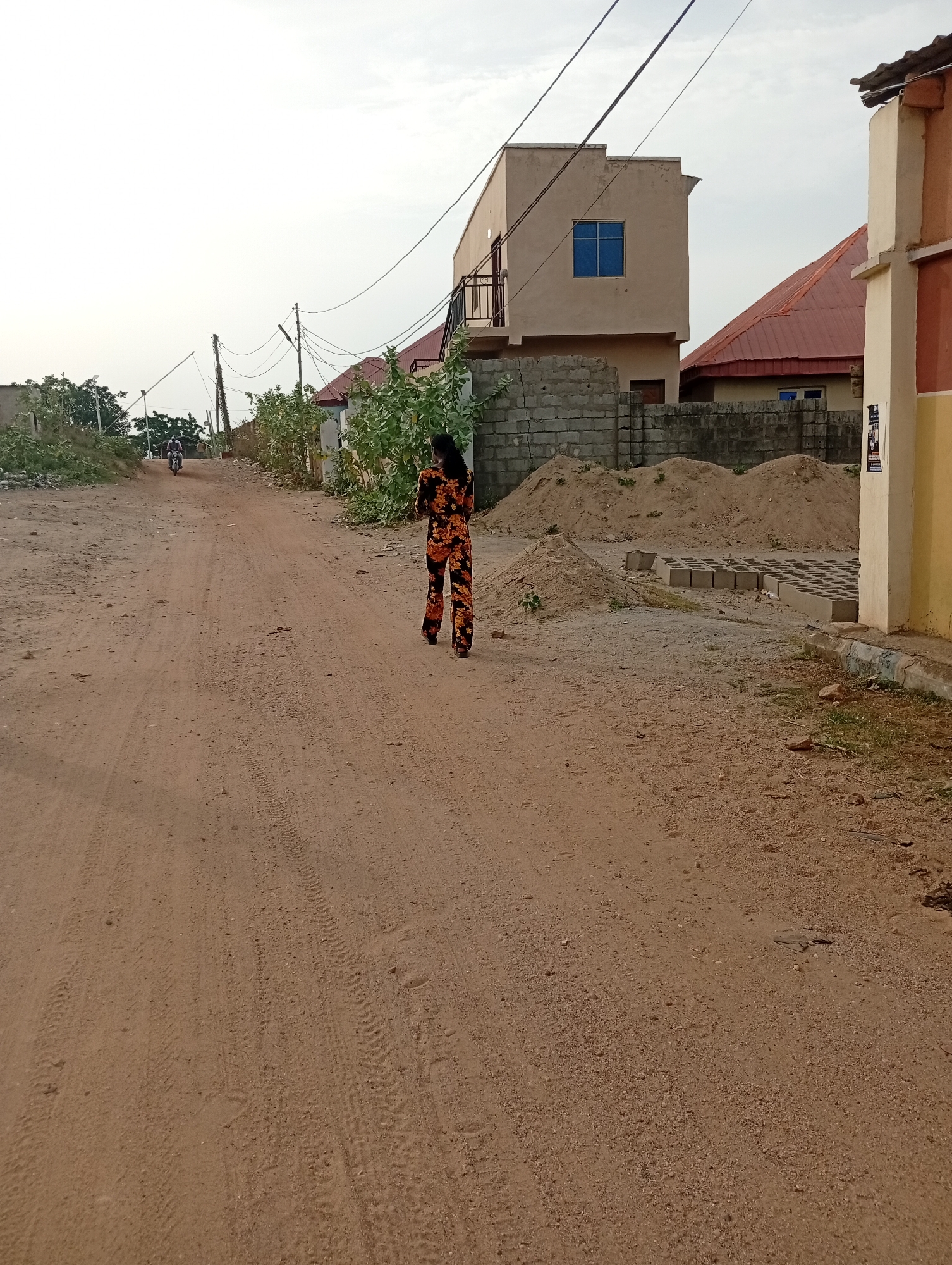 The image shows a dirt road in Adankolo, Nigeria. A woman in a patterned orange and black outfit is walking away from the camera. In the distance, a person is riding a motorcycle on the same road. To the right of the woman are cinder block walls and piles of sand, with stacked cinder blocks nearby. Buildings of various sizes and colors are visible in the background, along with overhead power lines. The sky is overcast.
