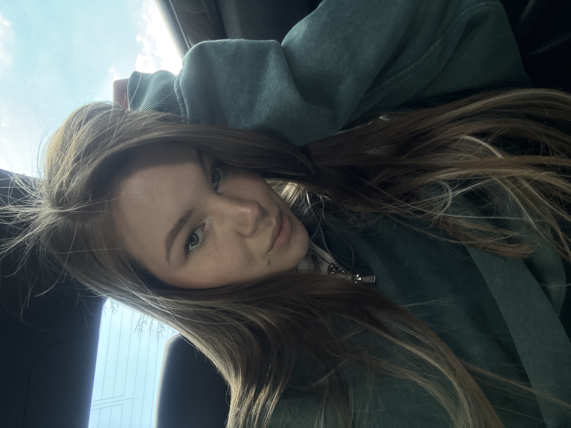 A young woman with long, light brown hair and light eyes is in a vehicle. She is wearing a green hooded sweatshirt or jacket. The background suggests she is near a window. The location cannot be identified as Belgrade, Serbia based on the given image.