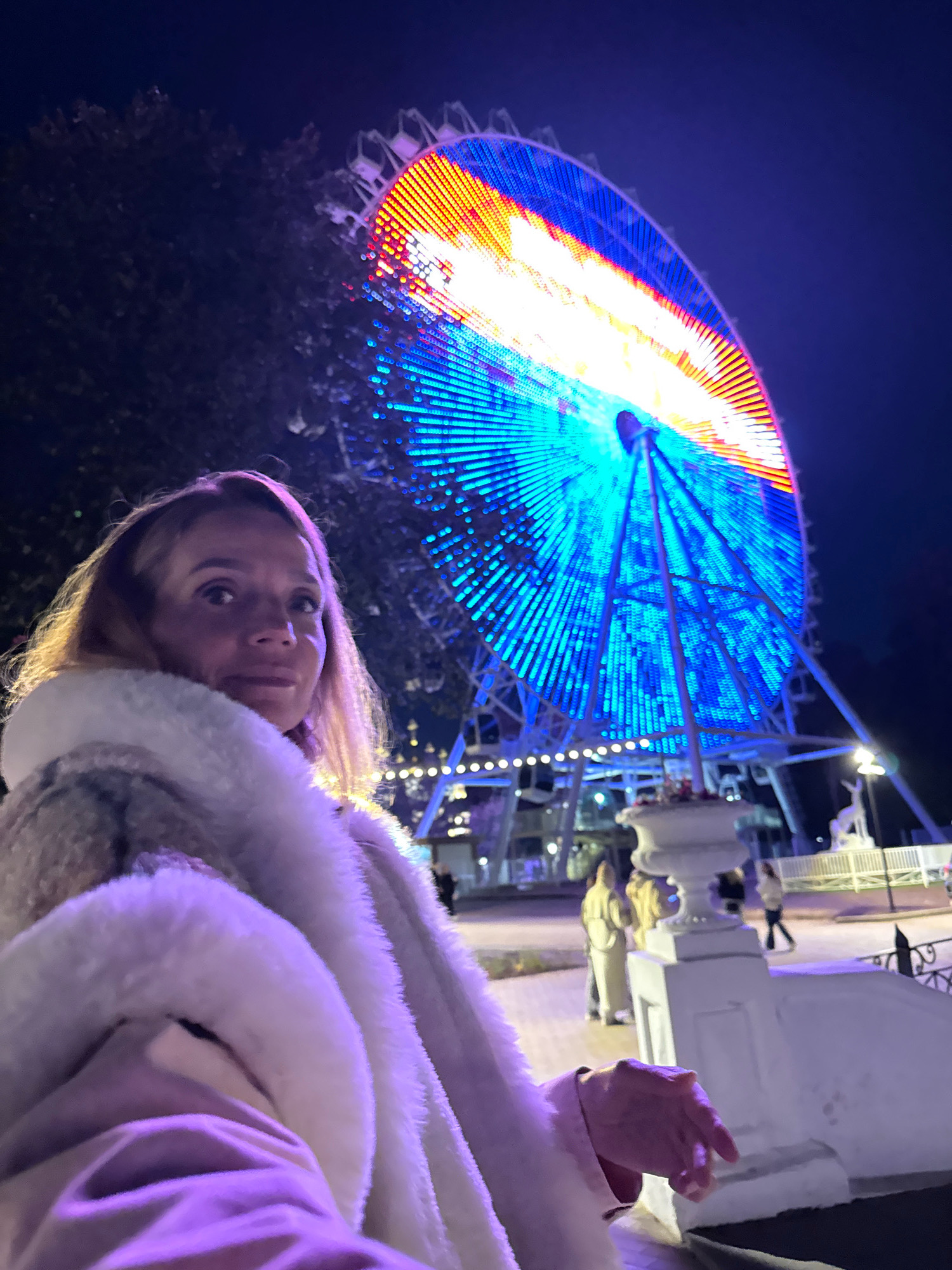 A medium shot captures a woman in the foreground, looking slightly off-camera to her right. She has blonde hair and is wearing a fluffy white coat. The background is dominated by a brightly lit Ferris wheel at night. The Ferris wheel displays a dynamic pattern of blue, orange, and white lights. In the midground, several people are visible walking, and there are structures that appear to be part of an amusement park or entertainment venue. The overall lighting suggests it is nighttime, with artificial lights from the Ferris wheel and other attractions illuminating the scene. The setting is the Tver Embankment in Tver, Russia.