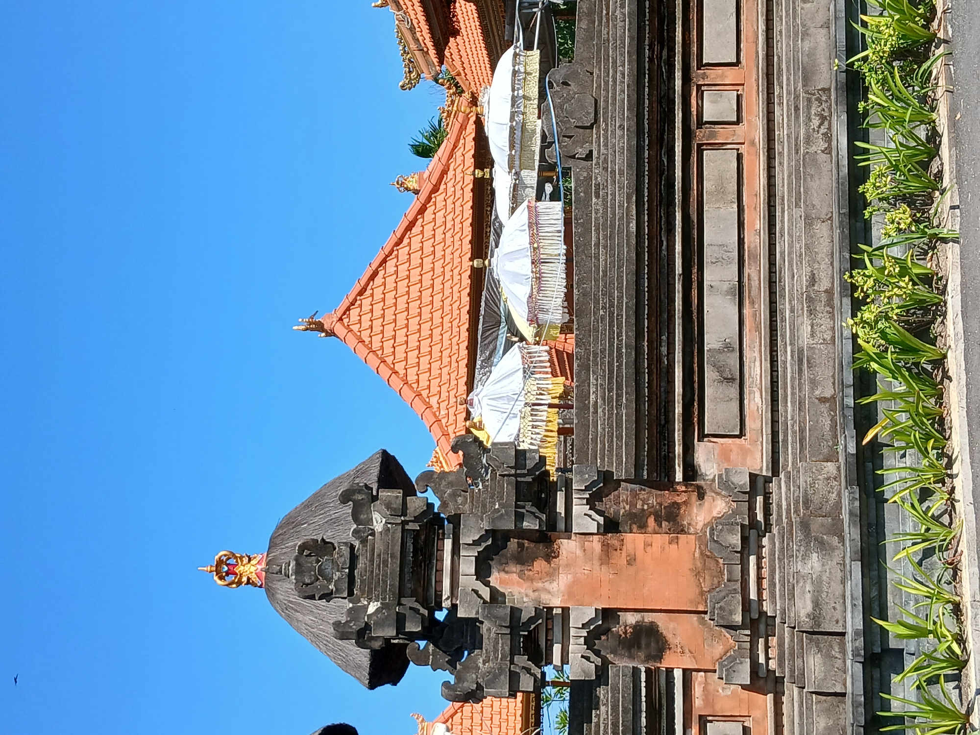 The image captures a section of the Pura Taman Ayun temple complex in Mengwi, Indonesia, under a clear blue sky. In the foreground, a stone wall with tiered levels separates the viewer from the temple grounds. The wall is constructed from grey stone blocks and features decorative elements. Above the wall, several ornate, white ceremonial umbrellas with gold fringe are displayed. These umbrellas are positioned in front of buildings with traditional orange tiled roofs. A prominent stone gateway structure, topped with a thatch-like roof and a golden crown ornament, stands to the left of the umbrellas. Green plants line the base of the wall in the foreground, and a bird is visible in the upper left corner of the sky. The architectural style is indicative of Balinese Hindu temples, characterized by intricate stone carvings, tiered roofs, and decorative elements like the umbrellas, often used in religious ceremonies. The scene is peaceful and conveys a sense of cultural and religious significance.