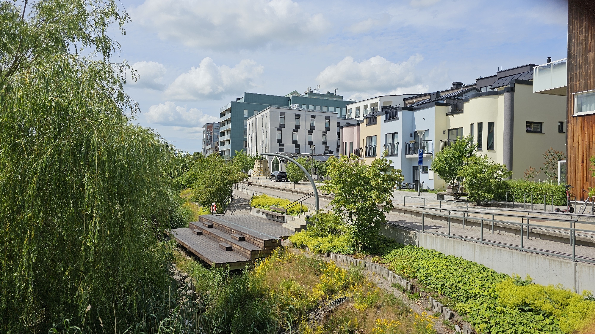 This image depicts a modern urban waterfront area in Linköping, Sweden, likely during daytime on a clear, partly cloudy day. In the foreground, lush green foliage from a weeping willow tree dominates the left side, with tall grasses and yellow wildflowers growing along a sloped embankment. A multi-tiered wooden seating area, resembling an amphitheater, is situated on this embankment, with a red life buoy hanging on a post to its left.
Further into the scene, a paved walkway runs parallel to a water feature or dry canal. Along this walkway, there are modern apartment buildings with diverse architectural styles and pastel-colored facades, including light blue, yellow, and cream. Some buildings have balconies with railings, and rooftop solar panels are visible on at least one structure. Benches are placed along the walkway, and there are several trees and green spaces interspersed between the buildings. A black car is parked near one of the buildings.
In the background, more apartment buildings of varying heights and colors can be seen, some with darker facades. The sky is predominantly blue with scattered white cumulus clouds. The overall impression is a well-maintained, contemporary residential area integrated with green spaces and recreational amenities.