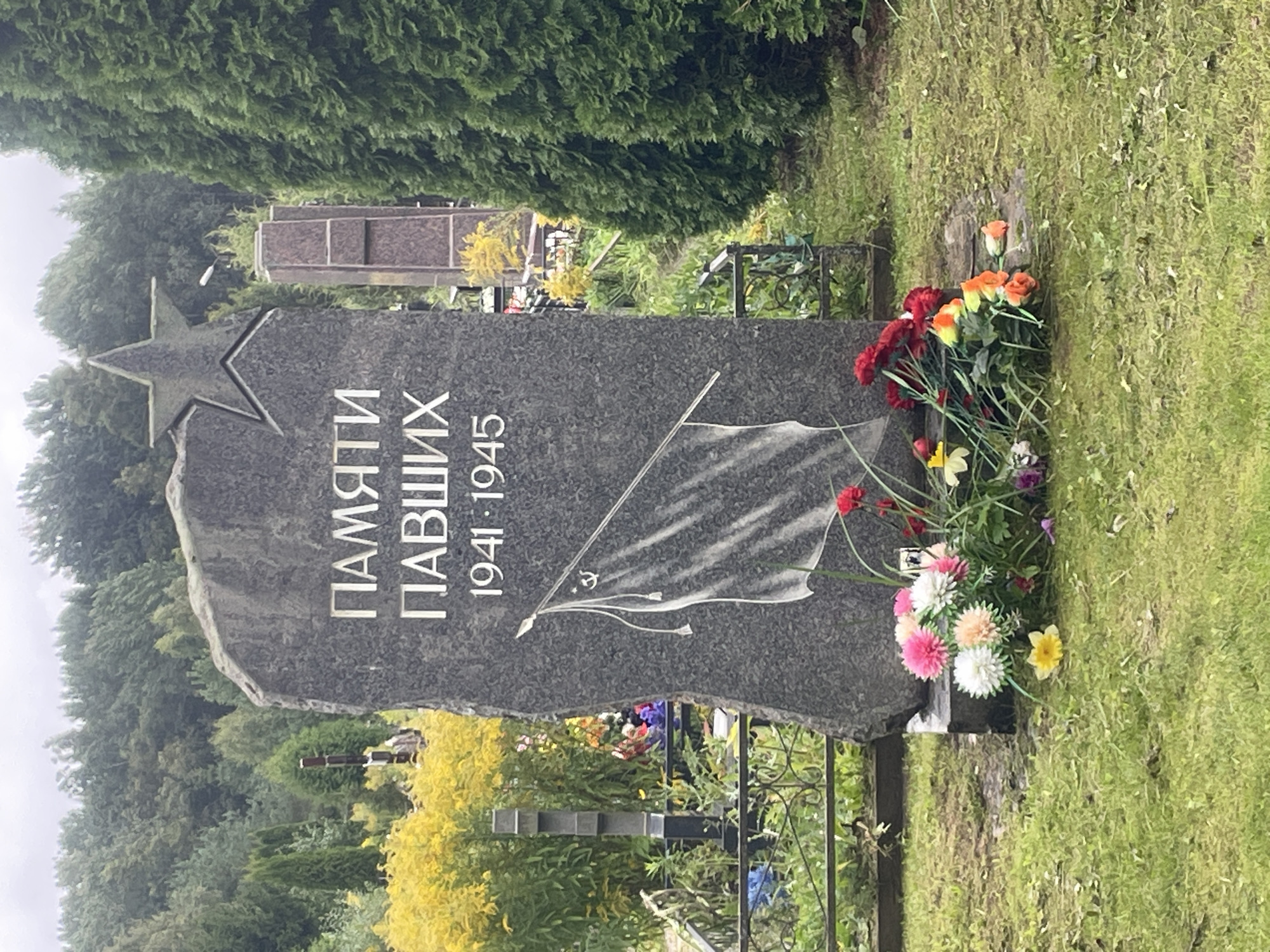 The image is a close-up, eye-level, full shot of a war memorial gravestone in Khimozi, Russia. The gravestone is made of dark gray granite and is carved into an irregular shape, with a five-pointed star at the top. White Cyrillic lettering is carved into the stone, reading "ПАМЯТИ ПАВШИХ 1941-1945" which translates to "In memory of the fallen 1941-1945". Below the inscription is a carved image of a flag on a pole with a sickle and hammer emblem. A cluster of artificial flowers, in various colors including pink, white, orange, and red, are placed at the base of the gravestone. One yellow artificial flower lies on the grass in front of the monument. The gravestone is situated on a grassy area, with a chain-link fence visible in the background. Behind the fence, there are more gravestones, some with crosses, and lush green trees. The overall environment suggests a cemetery.