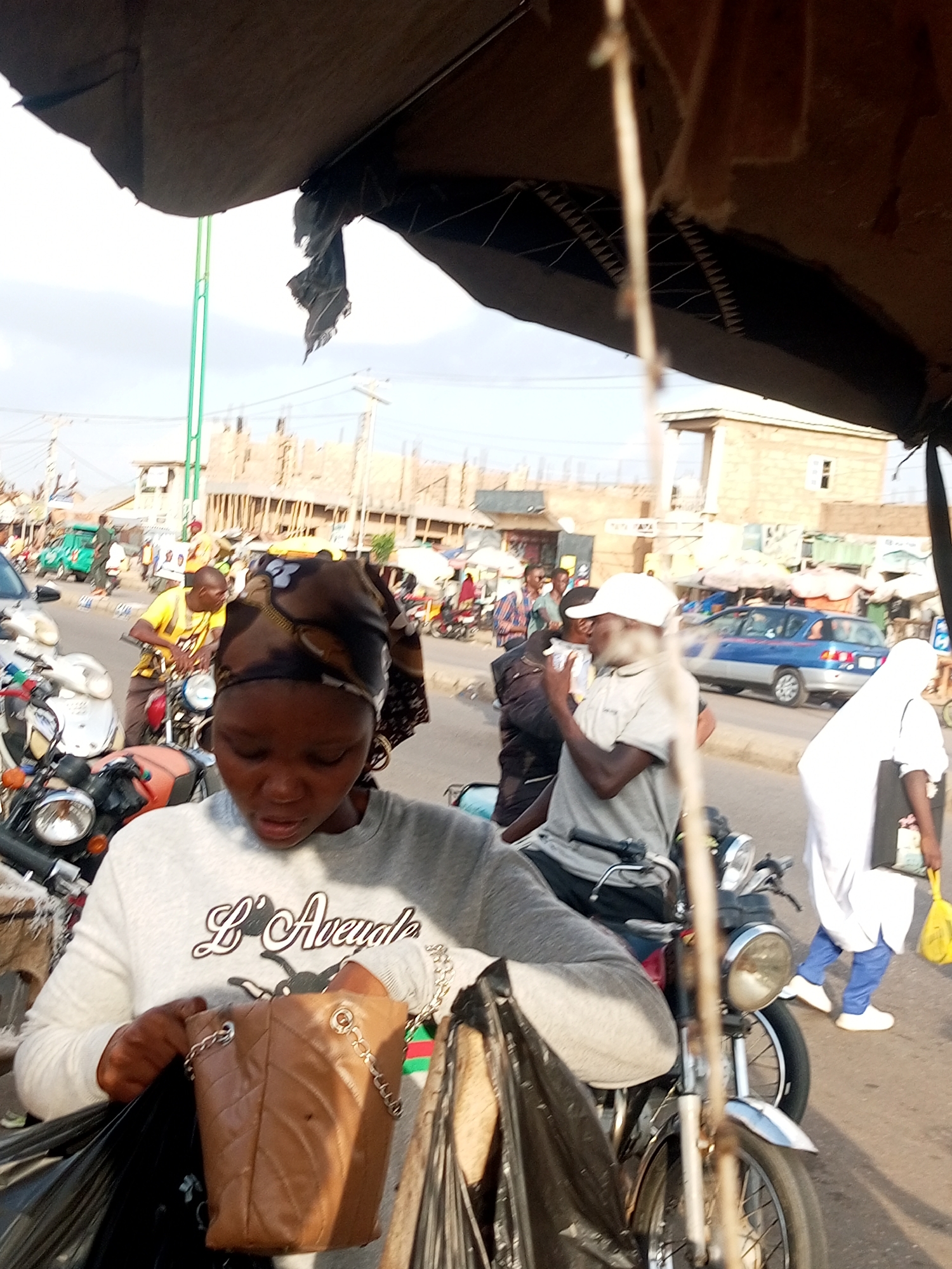 The image depicts a bustling street scene, likely in an urban environment, during daylight. The foreground is dominated by a young woman wearing a grey sweatshirt with the text "L'Avengele" printed on it, and a headscarf. She is holding a brown quilted handbag with a chain strap and appears to be interacting with black plastic bags, possibly packing or unpacking them. She is also partially sitting on a motorcycle.
In the background, several other individuals are visible. A man in a yellow t-shirt is on another motorcycle, and a person wearing a white cap and a light-colored shirt is also near motorcycles. Further in the background, there are buildings under construction, suggesting ongoing development. A blue car is driving on the road, and a person in a white outfit is walking away from the camera, carrying a yellow bag. The overall atmosphere appears to be that of a market or a busy thoroughfare with various modes of transportation.
The lighting suggests it is daytime, with bright sunlight casting shadows. The sky is visible and appears to be clear or partly cloudy. The presence of multiple motorcycles and the general activity of people suggest a lively and active setting, possibly a commercial area where people are shopping or commuting. The image does not provide explicit clues about the exact location beyond it being a street.