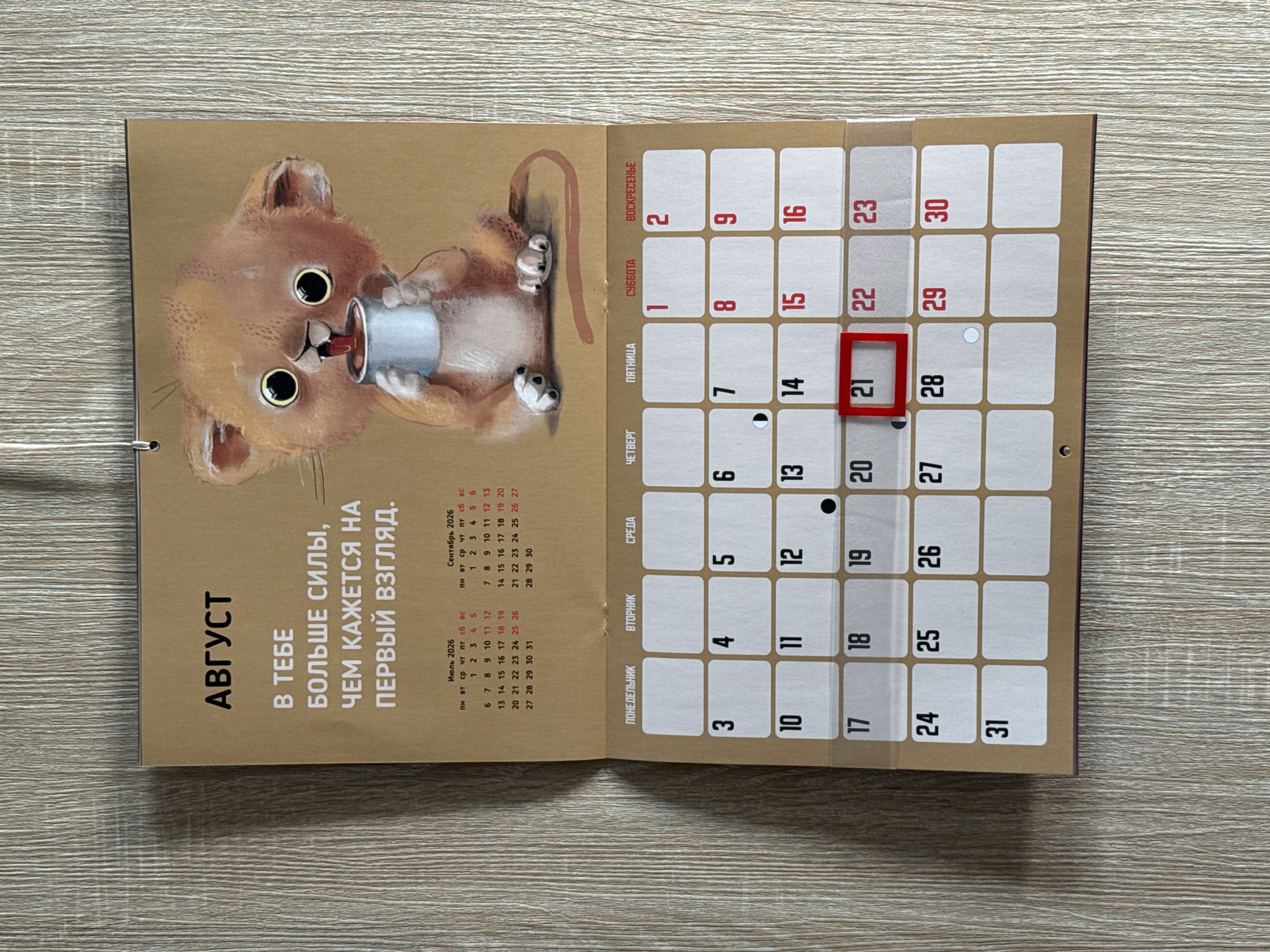 This is a close-up, eye-level, medium shot of a wall calendar depicting the month of August. The calendar is attached to a wall with a visible metal spiral binding at the top. The page features a cartoon illustration of a small, fluffy, lion-like cub holding a mug and drinking. Below the illustration, the month's title "АВГУСТ" (August) is displayed in large, bold, black lettering. Underneath the title is a motivational quote in Russian, "В ТЕБЕ БОЛЬШЕ СИЛЫ, ЧЕМ КАЖЕТСЯ НА ПЕРВЫЙ ВЗГЛЯД." (You have more strength than it seems at first glance.).
The calendar grid for August is displayed below the quote. The days of the week are labeled in Russian at the top of the grid: ПОНЕДЕЛЬНИК (Monday), ВТОРНИК (Tuesday), СРЕДА (Wednesday), ЧЕТВЕРГ (Thursday), ПЯТНИЦА (Friday), СУББОТА (Saturday), and ВОСКРЕСЕНЬЕ (Sunday). The dates are clearly visible within their respective cells. Notably, the 21st of August is highlighted with a red border, and the 28th has a small white circle. Small calendar grids for July and September 2026 are also visible at the top of the August page, providing context for the month's placement within the year.
The calendar is hung on a vertically textured wall that resembles wood paneling or a similar material. The lighting is even, and the image is in focus, allowing for clear visibility of all elements. The overall presentation suggests a domestic or office setting where this calendar is in use.
The provided image does not contain any information or visual cues that would allow for the determination of its location within the city of Lyubertsy, Russia.