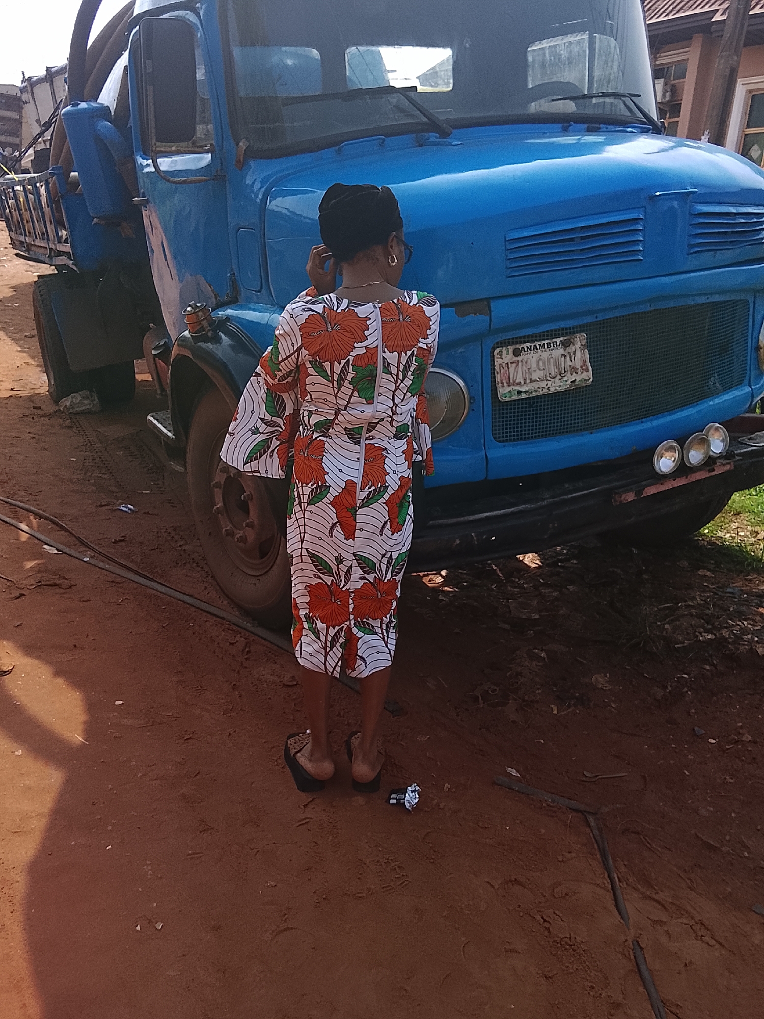 The image captures a full-length, rear view of a person standing on a reddish-brown dirt road, positioned in front of a large blue truck. The individual, likely a woman, is adorned in a vibrant, long-sleeved dress featuring striking orange and green floral patterns against a white background. She wears a black headscarf and black flip-flops, with gold hoop earrings visible. Her right hand is slightly raised towards her ear, suggesting she might be adjusting her hair, headscarf, or perhaps engaged in a phone call.
The prominent subject is a heavy-duty blue truck, which appears to be a tanker or a specialized cargo vehicle, indicated by the visible pipes and structures on its side. It is parked on the unpaved ground. The front of the truck displays a Nigerian license plate that clearly reads "ANAMBRA, Light of the Nation" and the registration number "NIZH-900XA." Three round auxiliary lights are mounted below the grille on the front bumper.
The setting is an outdoor area in Onitsha, Nigeria, characterized by uneven, reddish-brown soil, with visible tire tracks and sparse debris. Dark electrical cables or hoses lie across the foreground. A small piece of white discarded wrapper is seen near the woman's feet. The scene is illuminated by bright daylight, casting distinct shadows on the ground, indicating a sunny day. No specific activity or event is clearly unfolding beyond the woman's posture near the truck.