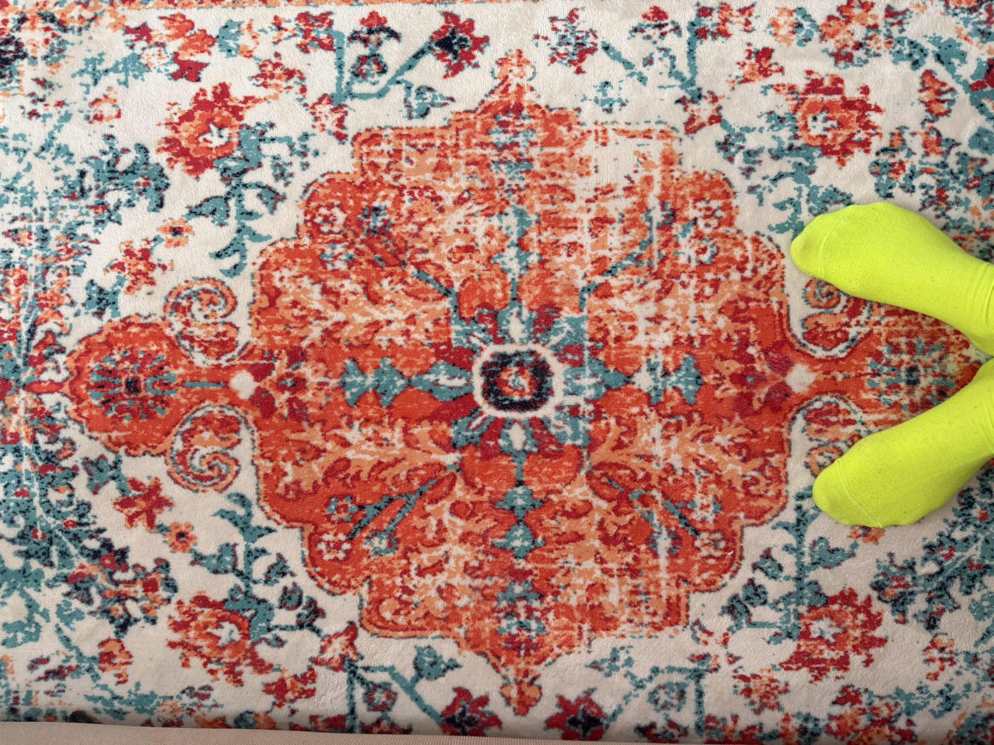 A close-up, aerial shot captures a section of a patterned rug with neon green socks resting on it. The rug has a white base with an ornate central design in varying shades of orange, red, and blue-green. The socks are situated on the right side of the rug, near the edge of the central pattern. The setting is unclear, and there is no discernible evidence to place this image in Petropavlovsk-Kamchatsky, Russia.