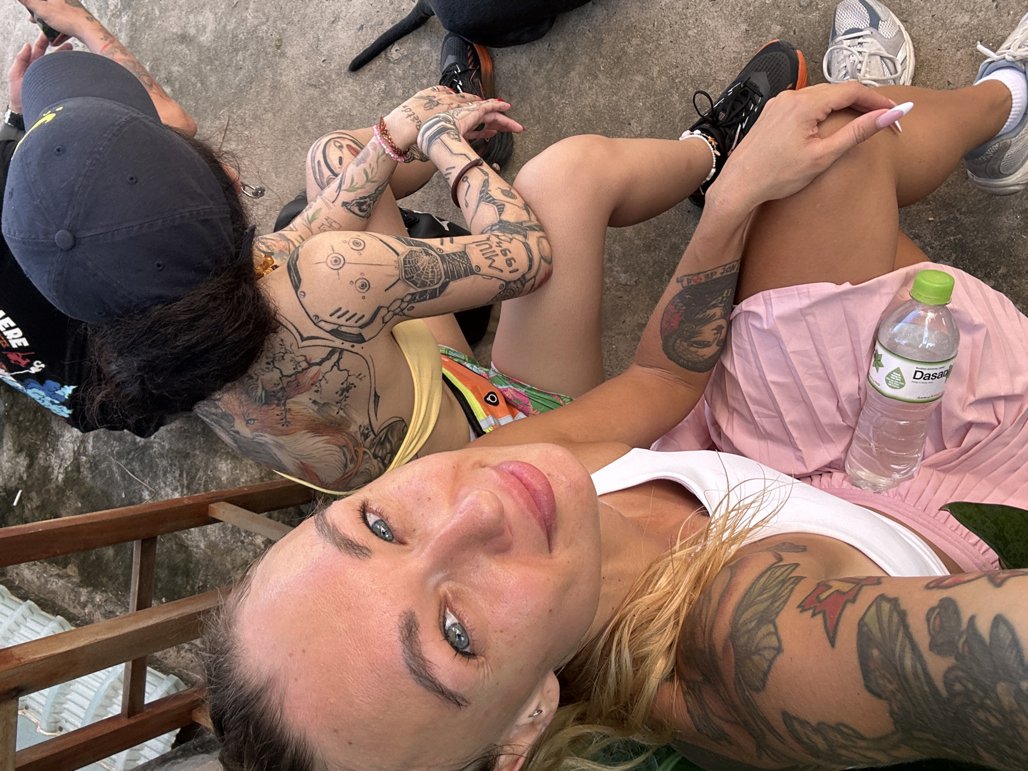 In Phu Thuy Ward, Vietnam, two individuals are seated outdoors on a concrete surface. The person in the foreground, a woman with blonde hair and multiple tattoos, is looking directly at the camera. She is wearing a white tank top and a pink pleated skirt. Beside her is a partially visible bottle of Dasani water. Behind her, another individual with dark hair and a black baseball cap is seated, also adorned with extensive tattoos on their arms and back. This person is wearing a yellow top and is looking down, potentially at a phone. The environment appears to be casual, with a wooden structure and corrugated roofing visible in the background.