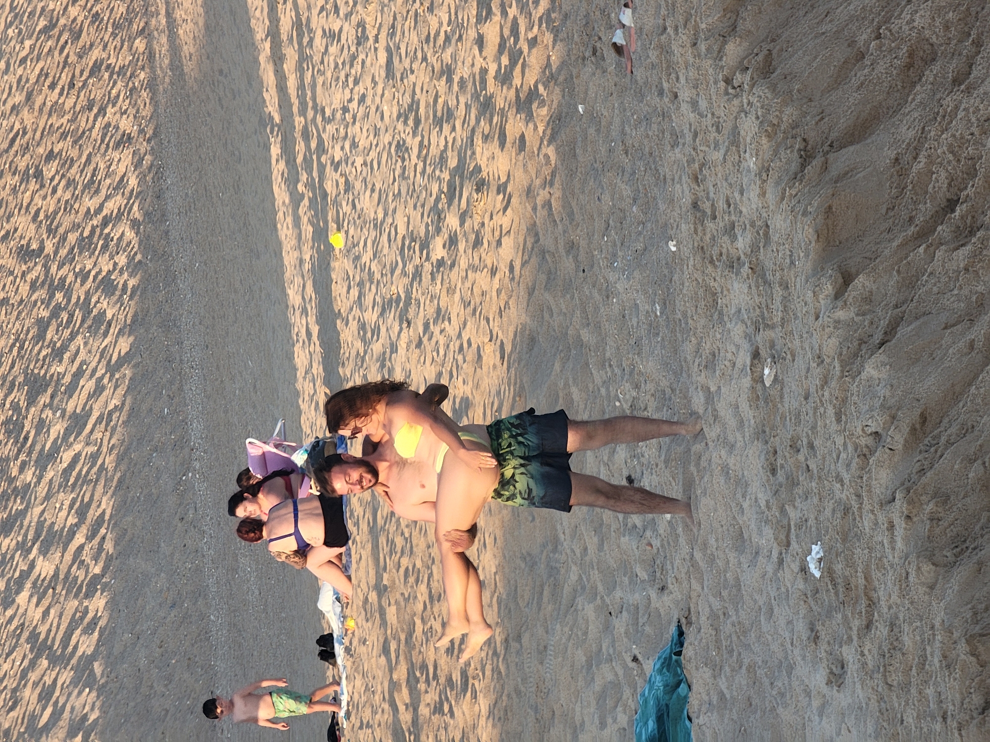 This is a high-angle, low-light, portrait-orientation photograph taken at Quarteira, Portugal, on a sandy beach. The scene is dominated by the texture of the sand, with ripples and shadows suggesting a late afternoon or early evening time. Several people are visible on the beach. In the foreground, a young boy in green swim trunks stands looking to the left. Further up the beach, a group of people are sitting and standing, some appearing to be on beach chairs. A couple is prominently featured in the center of the frame: a man in dark patterned swim trunks is carrying a woman wearing a yellow bikini. They are looking at each other and appear to be engaged in a playful or affectionate moment. The overall mood seems relaxed and casual, indicative of a beach outing. There are no visible signs of text in the image.