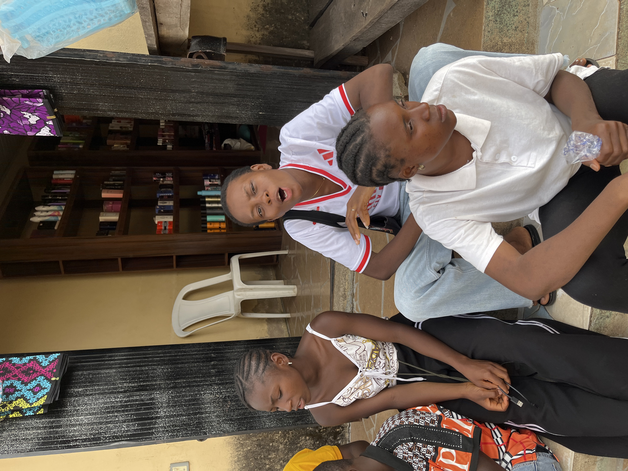 Three young women are in an outdoor area. One woman is sitting on another woman's lap. The woman sitting up is wearing a white shirt with red trim and has her mouth open. The woman she is sitting on is wearing a white button-down shirt. The third woman is sitting on the ground, wearing a white patterned top and black pants with white stripes. The location is suggested to be in Jos, Nigeria. A white plastic chair is behind the two women. A bookshelf and plastic-covered items are in the background.