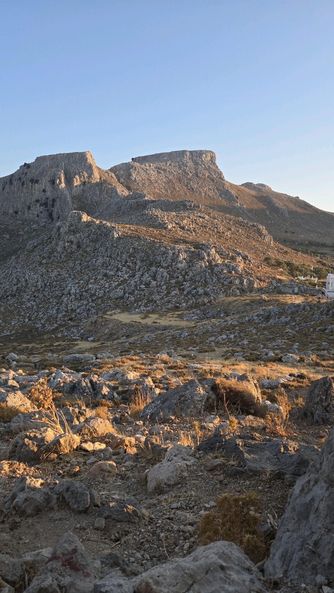 The image captures a rugged, rocky mountain landscape under a clear, pale blue sky, likely at sunrise or sunset given the warm, golden light illuminating the terrain. The foreground is dominated by a scattering of large, weathered grey rocks and dry, sparse vegetation. As the view recedes, the landscape transitions to a steeper, more imposing mountain range characterized by sharp, uneven rock formations and patches of dry grass and scrub. In the middle ground, nestled among the lower slopes, are a few white buildings, suggesting a settlement or isolated structures. A narrow, winding path is faintly visible in the rocky terrain. The overall impression is one of a remote, arid, and sun-drenched environment. This scene is likely located near the town of Archangelos, Rhodes, Greece, known for its mountainous and rocky terrain, and proximity to ancient sites and settlements.
