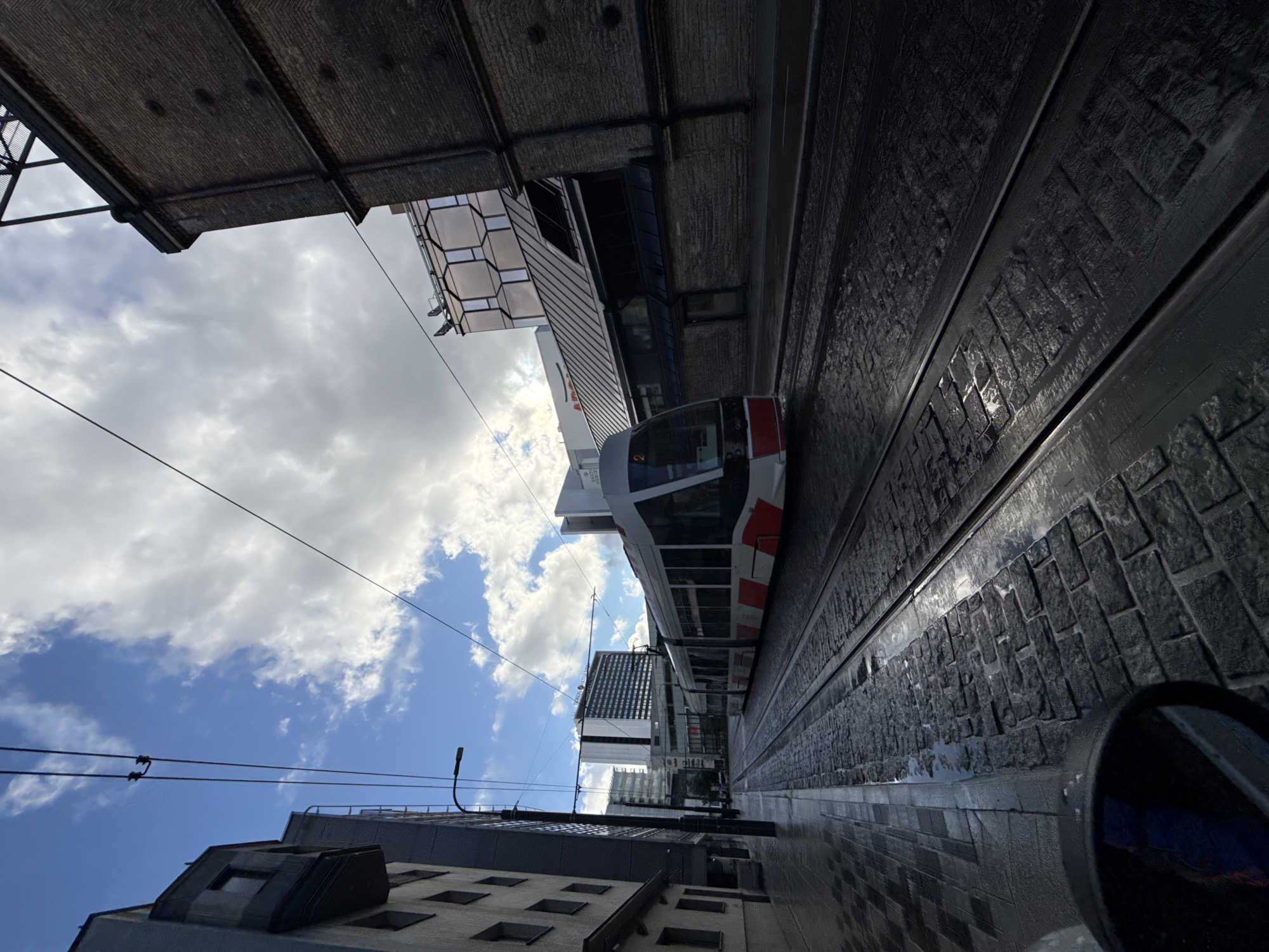 The image depicts a tram on a wet cobblestone street in Tallinn, Estonia. The tram, white with red accents, is moving towards the viewer on the right track. The street is lined with modern buildings and older brick structures. The sky is cloudy with patches of blue. The scene appears to be from a slightly elevated perspective, possibly from a vehicle.