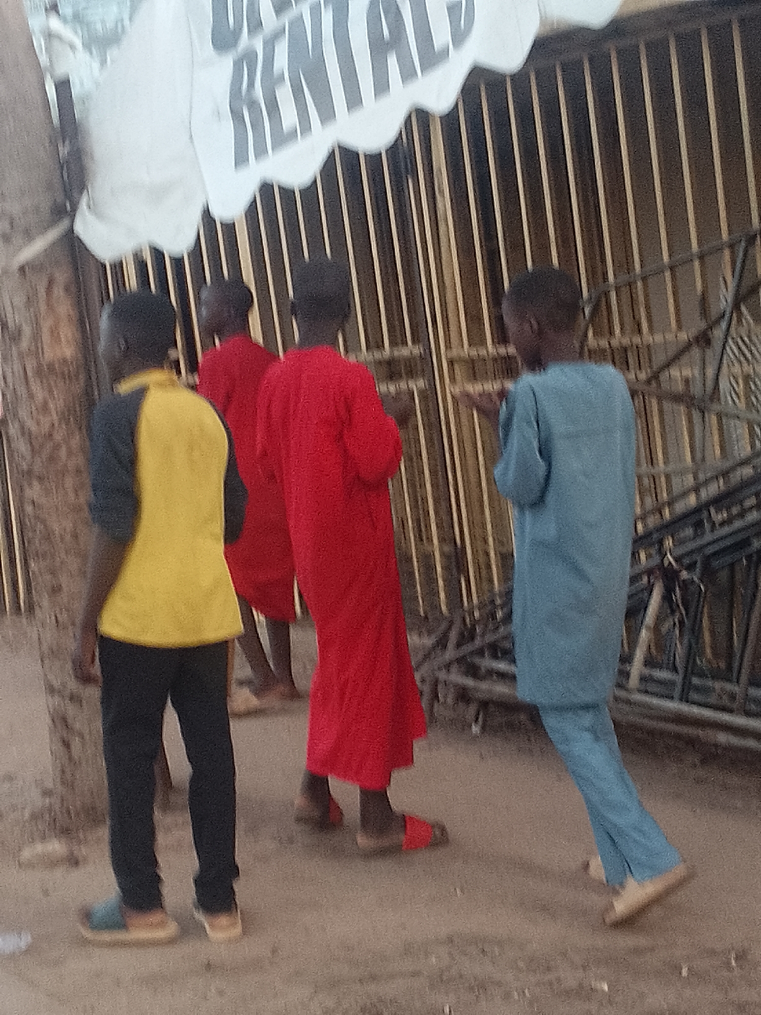 In Pantame, Nigeria, three young males are observed from behind standing on a dirt ground in front of a metal fence. The individual on the left wears a yellow shirt with dark sleeves and black pants, along with light blue and beige sandals. The middle individual is dressed in a long, bright red robe, and the person on the right wears a long, light blue robe. Behind the fence, a white banner with dark lettering is partially visible, with the word "RENTAL" discernible. The lighting suggests it is daytime, and the scene appears to be outdoors, possibly near a building or an establishment.