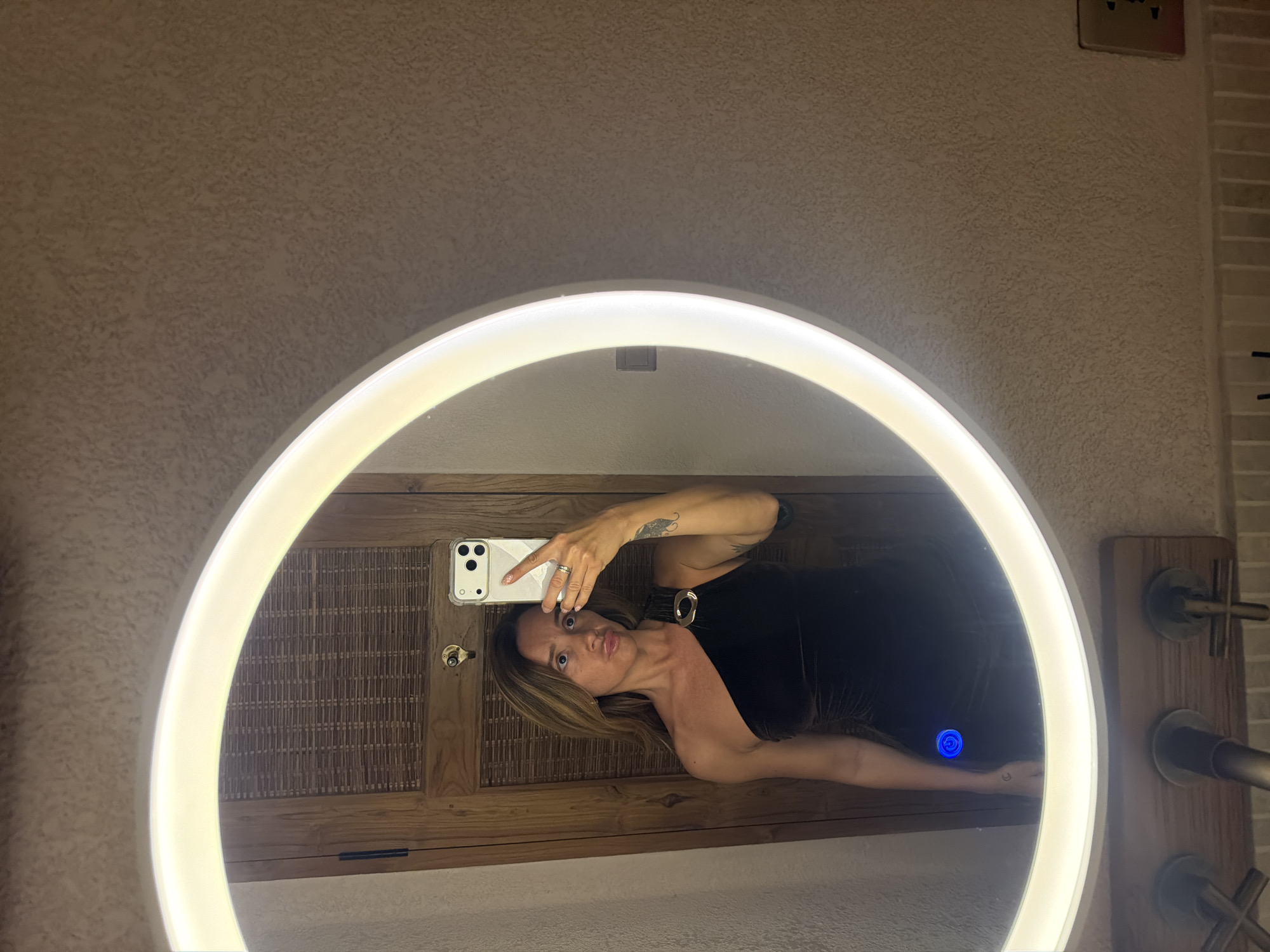 A woman with long blonde hair is taking a mirror selfie in a brightly lit bathroom. She is wearing a black one-shoulder dress with a gold buckle detail on the strap. Her left arm is raised to hold her phone, and her right arm is by her side. A tattoo of a crescent moon is visible on her left forearm. The mirror is round and has a built-in LED light. The background of the mirror reflects a wooden door with woven accents, a faucet, and a light switch. The bathroom walls appear to be textured. The image appears to be taken in the city of Mengwi, Indonesia.