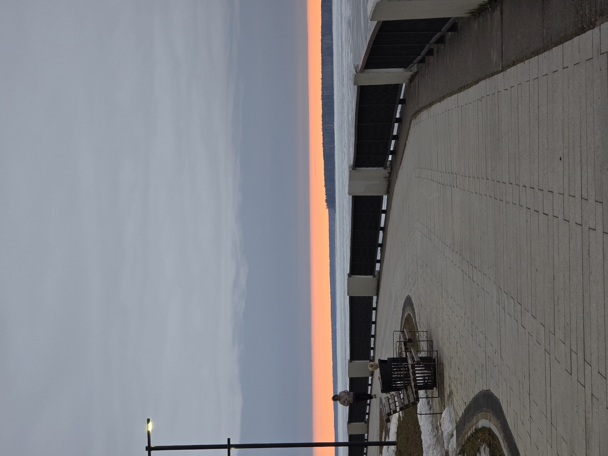The image shows an embankment view in the city of Chaikovsky, Russia, at what appears to be sunset. To the left of the embankment is a body of water with a strip of trees along the horizon. Above the water is a gray sky with hints of pink. The embankment is constructed of light-colored stone blocks. A black metal fence borders the water, connected by white concrete columns. Adjacent to the fence is a gray curb. A man in light-colored clothing stands near a bench, and snow patches are seen near the bench. A light post is visible in the foreground, with the light on.