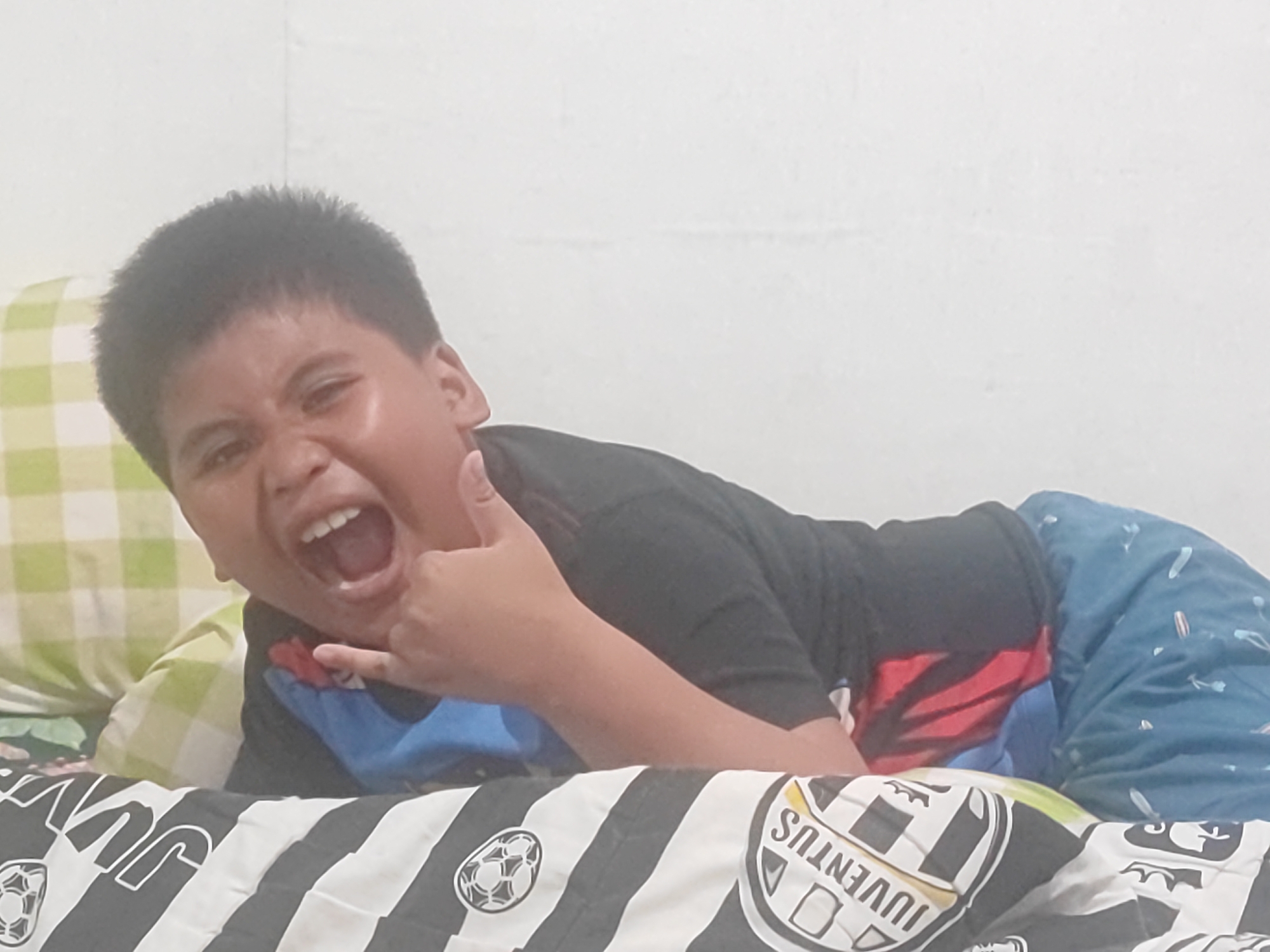 A young boy, appearing to be around 8-10 years old, is lying on a bed and making a rock-and-roll hand gesture with his right hand. He has dark hair and is wearing a black t-shirt and blue shorts with a patterned design. His mouth is open in a wide expression, and he seems to be looking towards the camera. The bed has a black and white striped comforter with what appears to be a soccer ball pattern and the word "JUVENTUS" printed on it. In the background, a light-colored wall is visible, and to the left of the boy, a pillow with a green and white checkered pattern can be seen. The overall setting suggests a bedroom. The image was taken in Tenggarong, Indonesia.