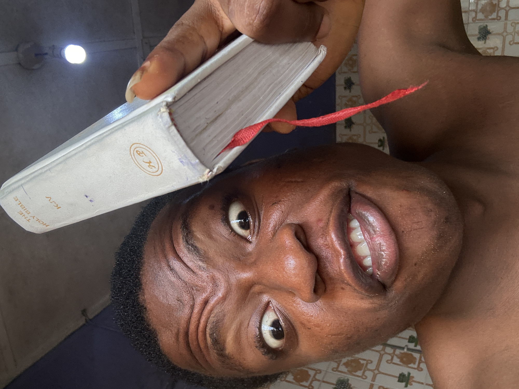 A close-up selfie shows a person holding a book above their head. The book appears to be a white-covered edition of "The Holy Bible KJV" with a red ribbon bookmark. The person's face is prominent, with visible skin texture and expression. The background includes a section of a tiled surface and a ceiling fixture. The location is not identifiable from the image.