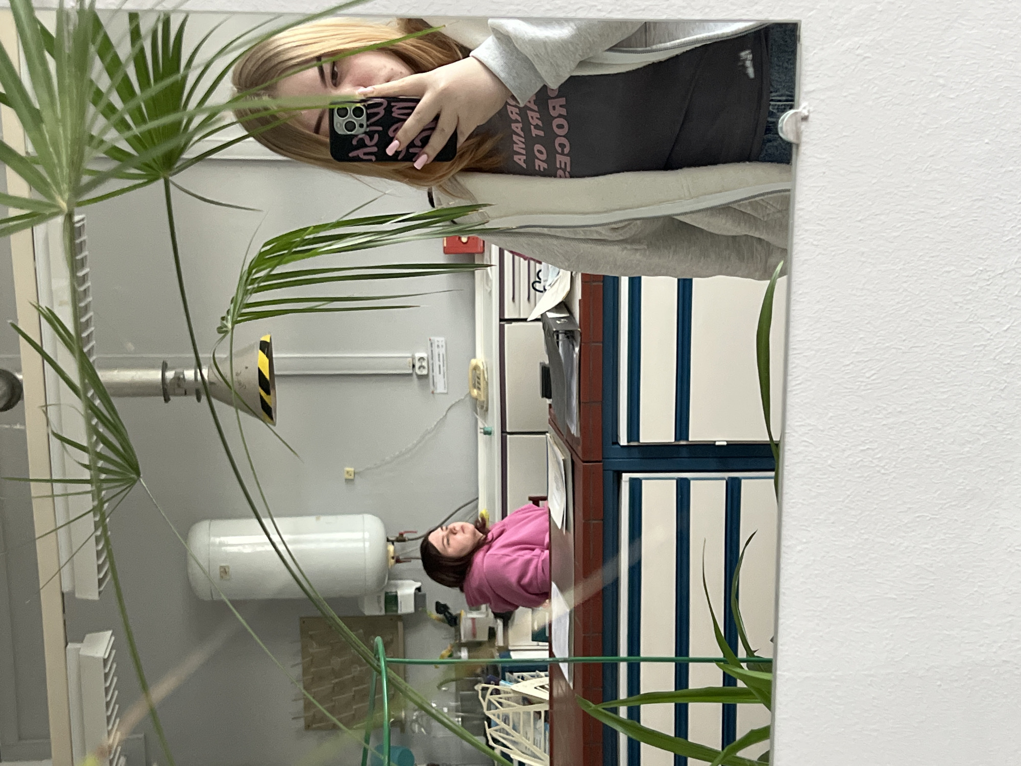A reflection in a mirror shows two women in a laboratory or classroom setting. One woman, facing the mirror, holds a phone with a black and patterned case up to take a picture, partially obscuring her face. She has blonde hair, pink nails, and is wearing a gray hoodie over a dark-colored top with pink text. The reflection reveals another woman, wearing a pink top, sitting at a desk in the background. The room appears to be a lab or classroom with standard equipment such as counters, cabinets with white and blue striped doors, and a large white cylindrical object mounted on the wall. A potted plant with green foliage is positioned in the foreground, partially obscuring the view. The wall adjacent to the mirror is textured and painted white. The location is Chaikovsky, Russia.