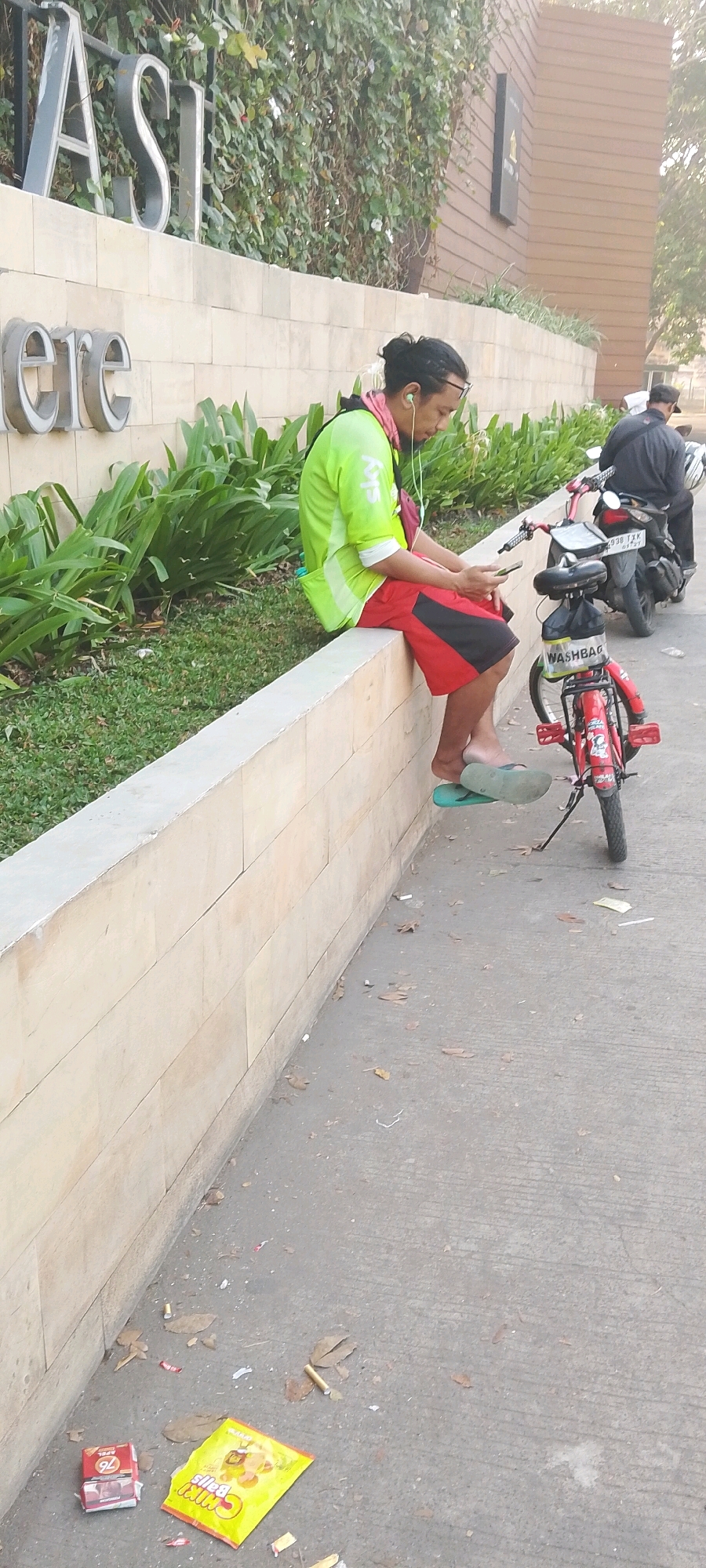 The image depicts an outdoor scene in front of a building with large metallic letters spelling "ASI" above smaller metallic letters spelling "ere" on a stone wall. A man is seated on a low, beige stone wall, wearing a bright green shirt with "sky" printed on it, red shorts with black accents, and flip-flops. He has earphones in and is looking at his mobile phone. A small red bicycle with a "WASHBAG" label on it is parked next to him. Further in the background, another person is seated on a motorcycle. The ground is paved with concrete and littered with fallen leaves and cigarette butts. To the left of the scene, a packet of "Chiki Balls" snacks and a box of what appears to be cigarettes are on the ground. The environment suggests a public space, possibly a commercial or residential complex entrance in Satriamekar, Indonesia.