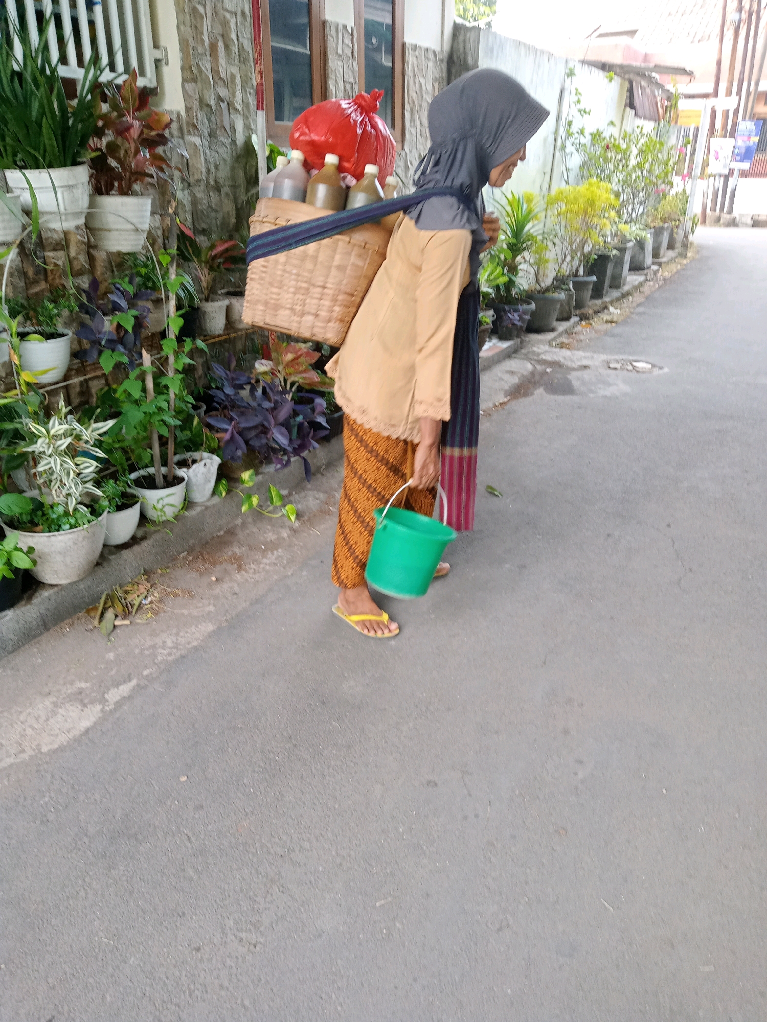 In Cirebon, Indonesia, a person wearing a grey hijab and a tan top, stands on a paved road. They are carrying a woven basket on their back, filled with bottles and a red plastic bag. In their right hand, they hold a green bucket. The person is wearing orange and brown patterned pants and yellow flip-flops. They are positioned next to a wall with potted plants of various sizes lining the walkway. The background shows more potted plants and a building.