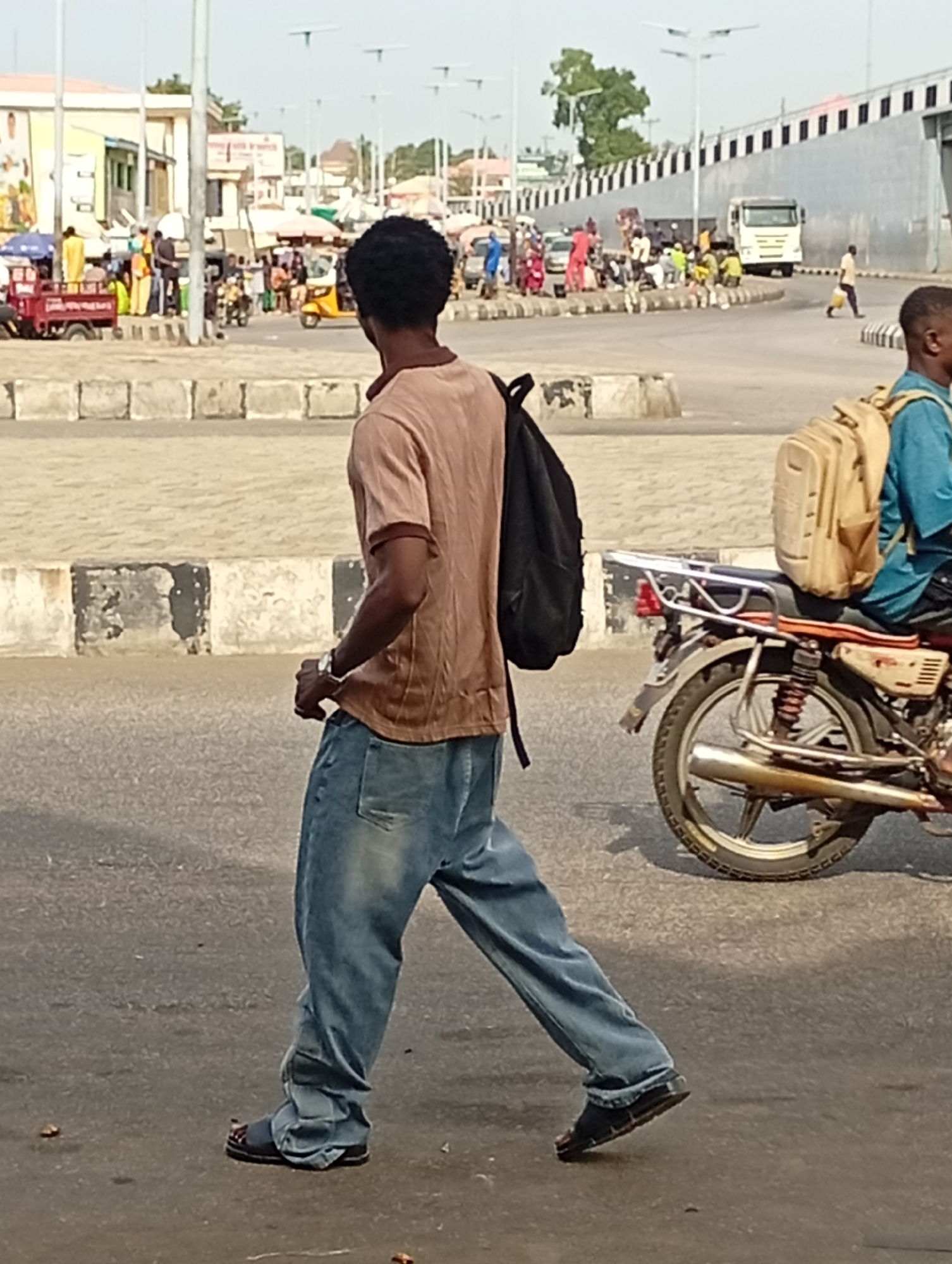 A medium shot shows a man crossing a street in Lokoja, Nigeria, with a black backpack slung over his left shoulder. He wears a light brown t-shirt and faded blue jeans, with dark sandals. A motorcycle with two riders, one wearing a blue shirt and a tan backpack, is partially visible to the right. In the background, across the street, there's a bustling street scene with people, parked vehicles like tuk-tuks and cars, and market stalls near buildings. Above the street, a white concrete wall stretches into the distance. The sky is overcast.