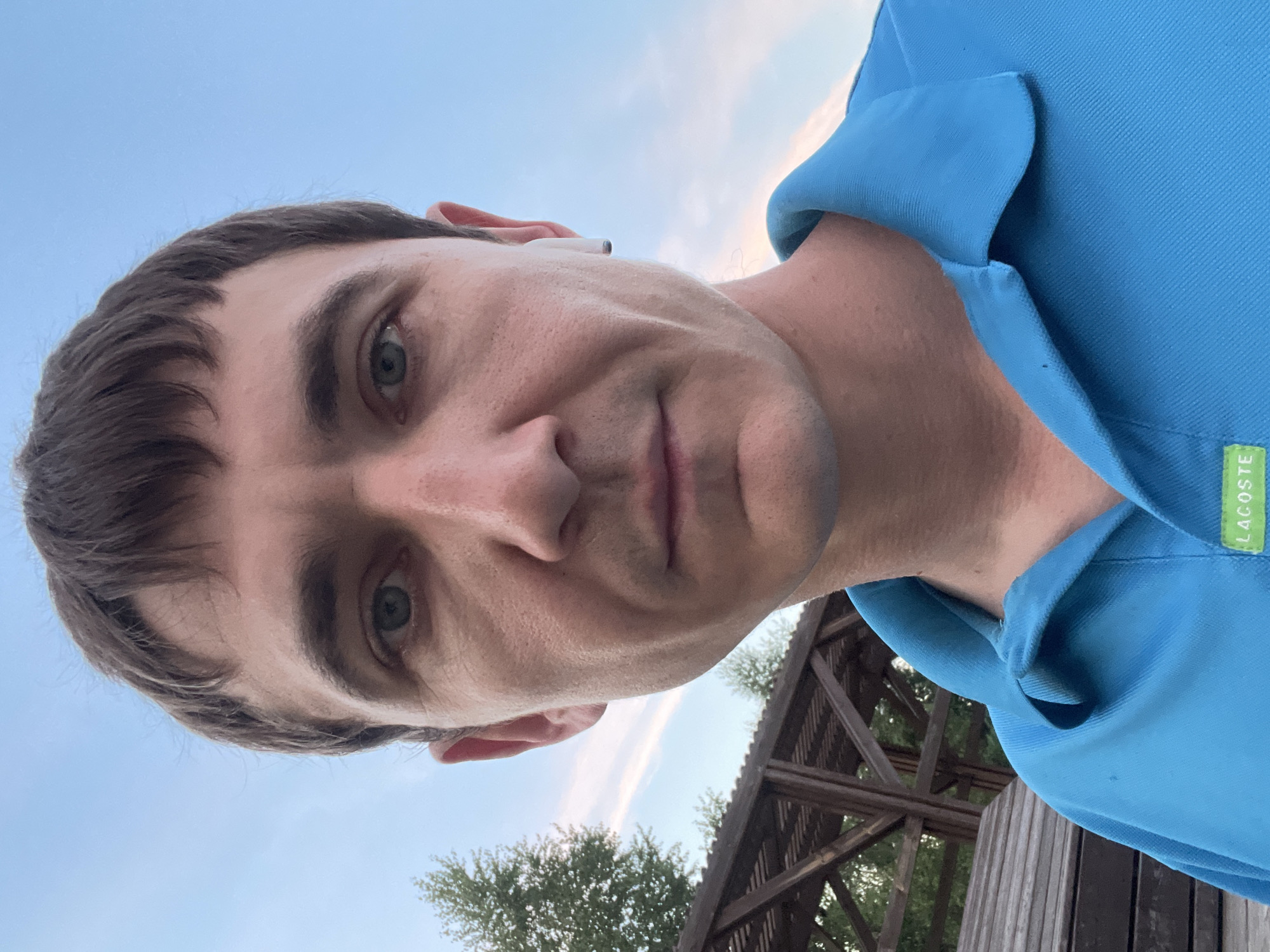 A close-up, outdoor, selfie-style photograph features a Caucasian man with short, dark brown hair and light blue eyes. He is wearing a bright blue collared polo shirt, which has a green rectangular tag with white lettering that reads "LACOSTE" visible on the lower right side. He has a white wireless earbud in his right ear. The man is looking directly at the camera with a neutral expression.
The background includes a clear, light blue sky with some wispy white clouds. To the left of the man, there is a wooden structure, possibly a pergola or a gazebo, with horizontal slatted roofing and vertical supports. Adjacent to the structure is a wooden bench or seating area. In the upper left portion of the image, the tops of several green trees are visible. The overall setting appears to be a park or an outdoor recreational area. The photograph was taken in Laishevo, Russia.