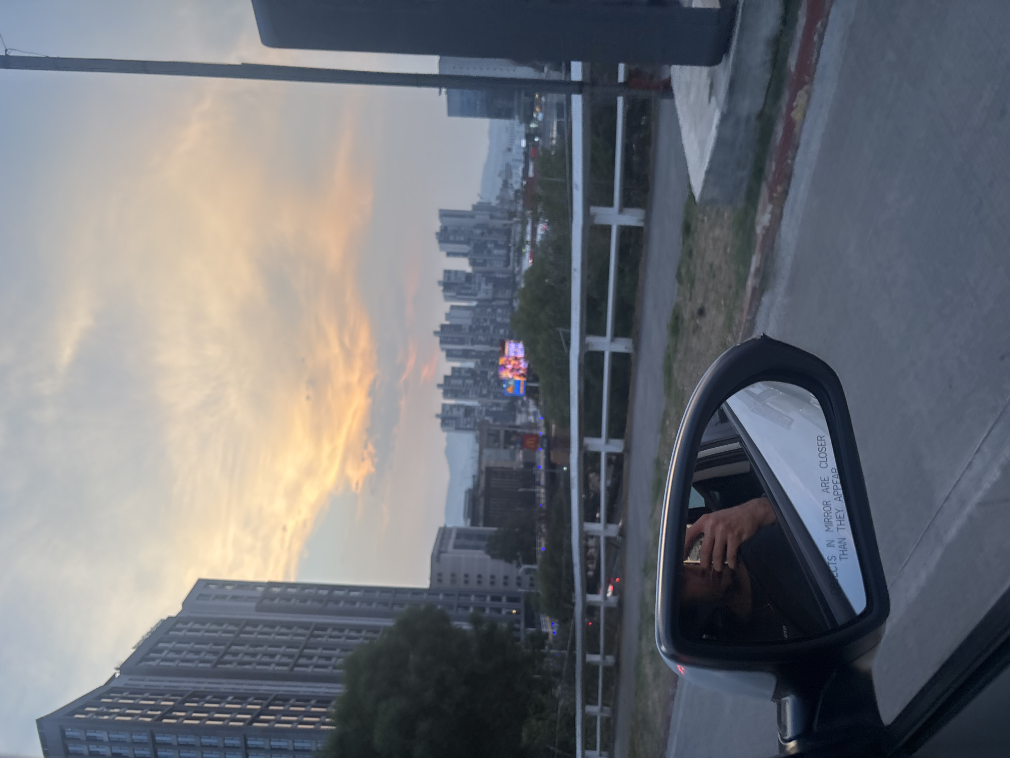 The image, taken from inside a vehicle, shows a cityscape at dusk, likely in Huixquilucan de Degollado, Mexico, given the urban density. The buildings, including a high-rise, are silhouetted against the fading light and orange-tinged clouds. The image is framed by the vehicle's side mirror, reflecting the photographer taking the picture with a phone. The vehicle appears to be on a road with a concrete barrier and vegetation.
