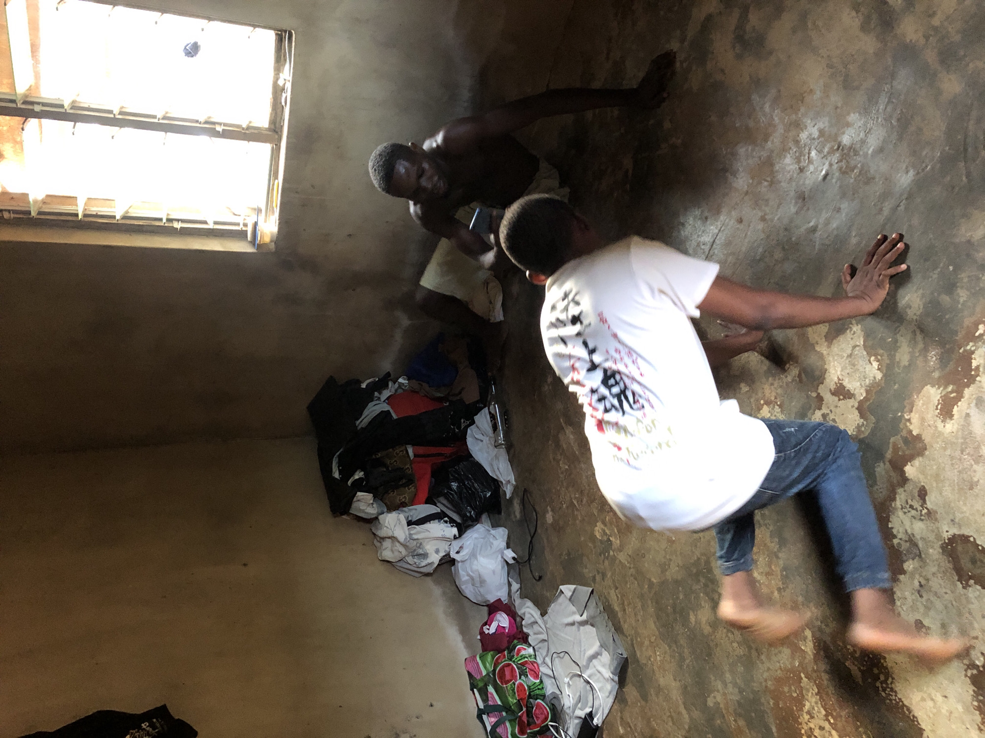 Two men are in a room with bare walls and a window. One man is fair-skinned, wearing a white t-shirt and blue jeans, and appears to be using his hands to brace himself against the wall. The other man is dark-skinned, shirtless, and holding a phone. A pile of clothing is on the floor. Location is Unknown, Unknown.