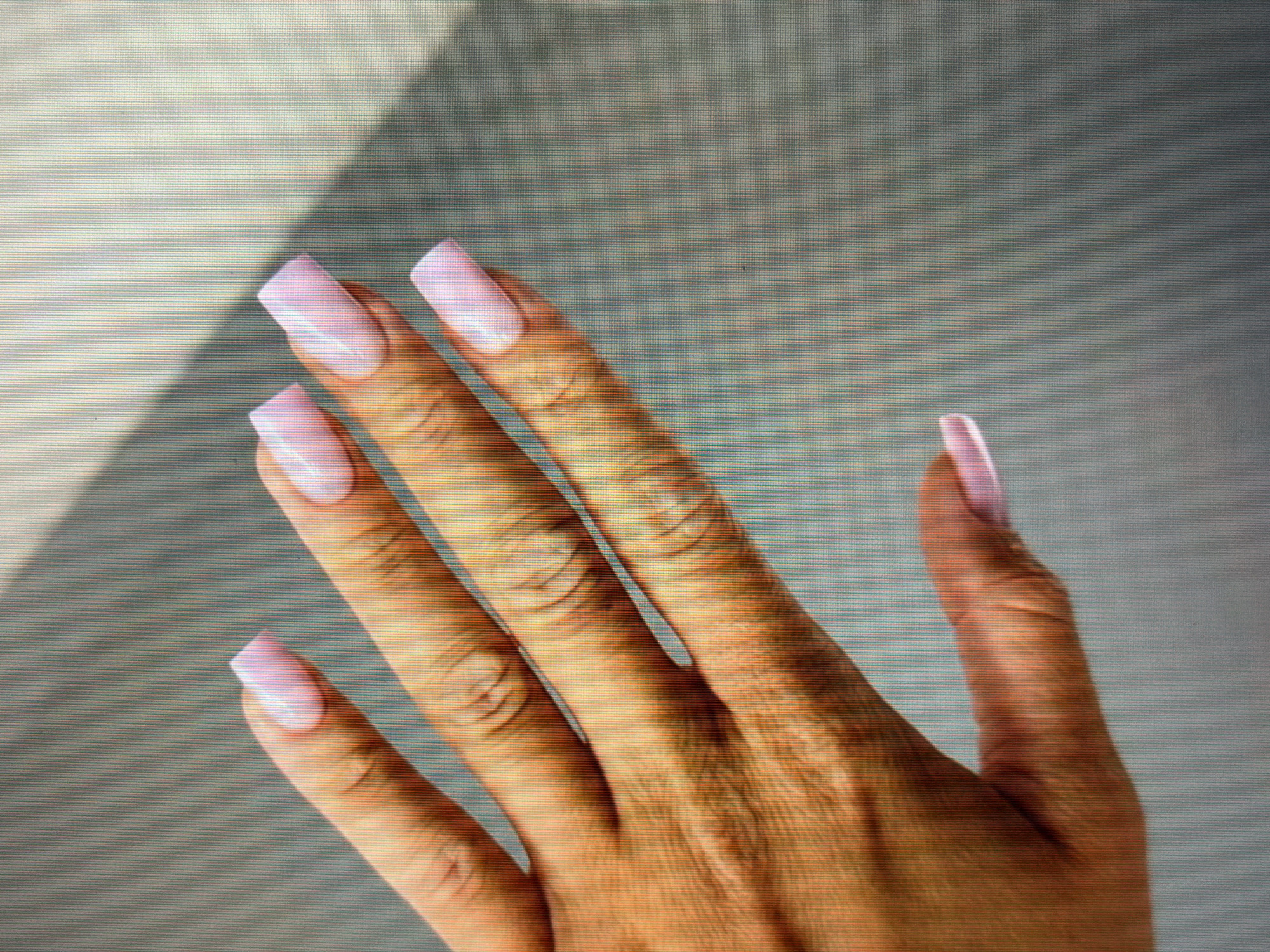 The image shows a close-up of a person's hand with light pink, square-shaped acrylic nails. The hand is positioned against a blurred background with diagonal shadows. There are no visible identifying features or environmental context that would suggest a specific location within Sirius, Russia, or any other city. The focus is solely on the hand and the nails.