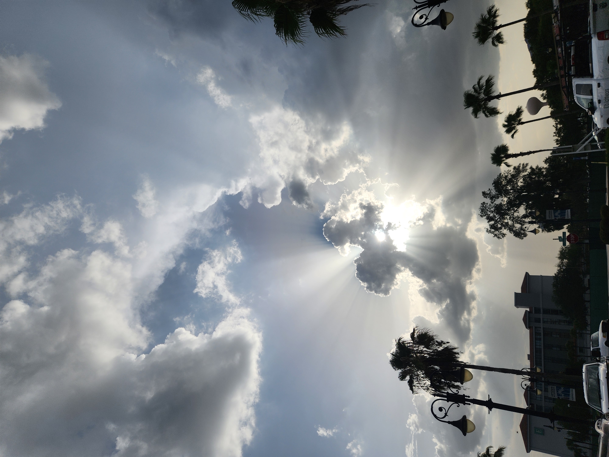 The image depicts a dramatic sky with sunbeams piercing through a dense cloud formation, creating a radiant effect. The scene is set in Altamonte Springs, Florida, as indicated by the presence of palm trees and a building with a sign that says "UPTOWN ALTAMONTE". In the foreground, a white pickup truck is parked, and the silhouettes of lampposts and other trees are visible against the bright sky. The overall atmosphere is one of a partly cloudy day with a strong emphasis on the celestial display.