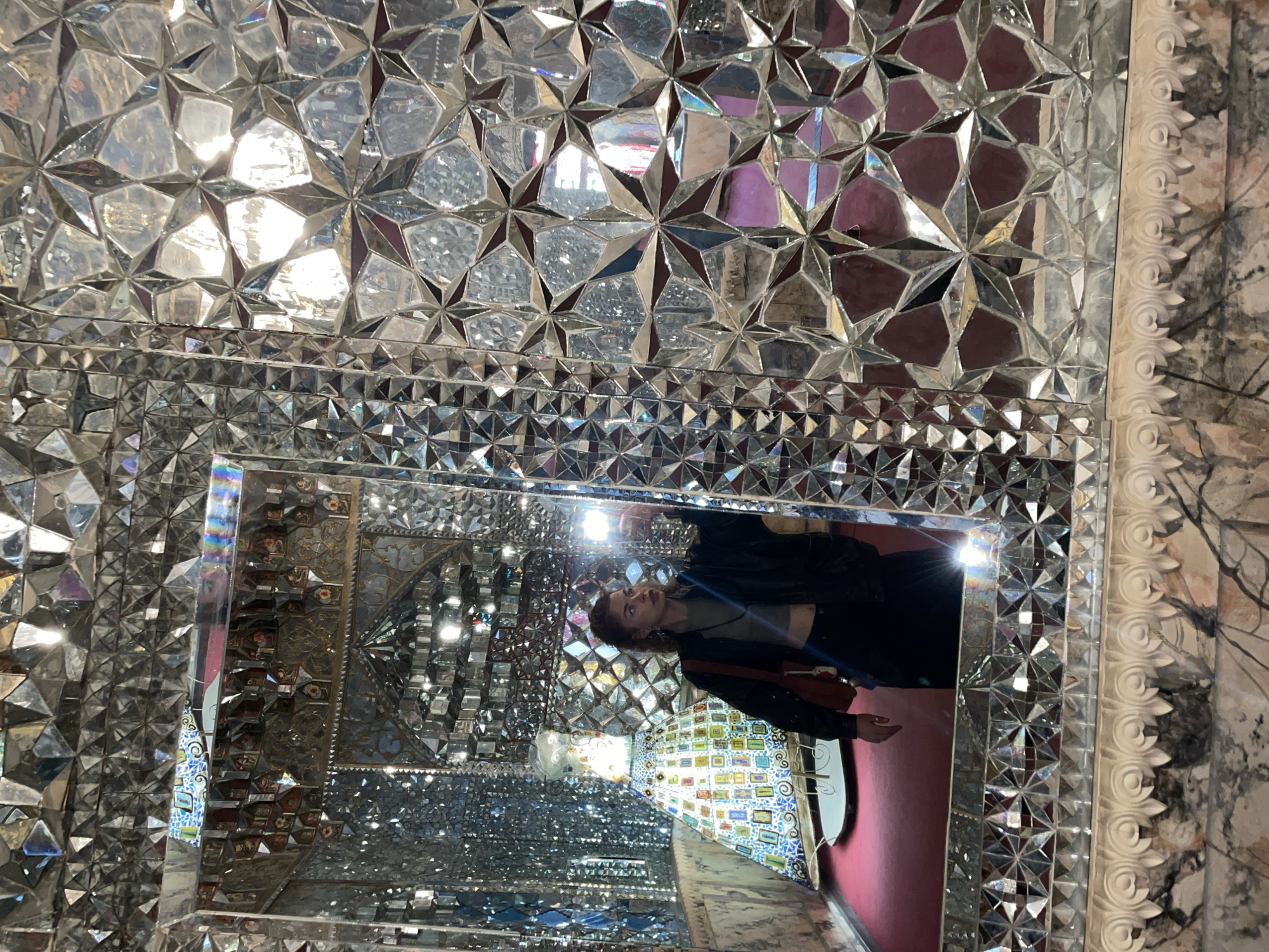 The image captures a woman reflected within an ornate, kaleidoscopic mirrored room, giving a sense of infinite reflections and fragmented light. The photograph is oriented vertically, showing the woman sideways, suggesting she is standing upright.
The main subject is a woman, likely in her 20s or 30s, with light brown hair, wearing a dark jacket, a light-colored crop top, and dark pants. She carries a reddish-brown shoulder bag. Her gaze is directed towards the camera or her own reflection, with a neutral expression.
The setting is an elaborate indoor space, possibly a hall or corridor, completely covered in intricate mirror work. The surfaces are composed of countless small, faceted mirror pieces arranged in geometric patterns, primarily stars and triangles, creating a dazzling and complex visual effect. The reflections show parts of the room: a red carpet or floor, and another wall adorned with colorful mosaic tiles, possibly depicting small floral or abstract patterns. A decorative white carved frame, with a scroll and leaf motif, runs along the right edge of the central mirror, separating it from a textured, possibly marbled wall section.
The room is brightly lit, with strong light sources causing highlights and glares across the mirrored surfaces, enhancing the shimmering, fragmented appearance. The continuous reflections create a disorienting yet captivating illusion of depth. The woman appears to be observing her surroundings or posing for a photograph in this visually striking environment, possibly a tourist attraction or an art installation. The location context suggests this unique mirrored room is found in Tbilisi, Georgia. There is no visible text in the image.