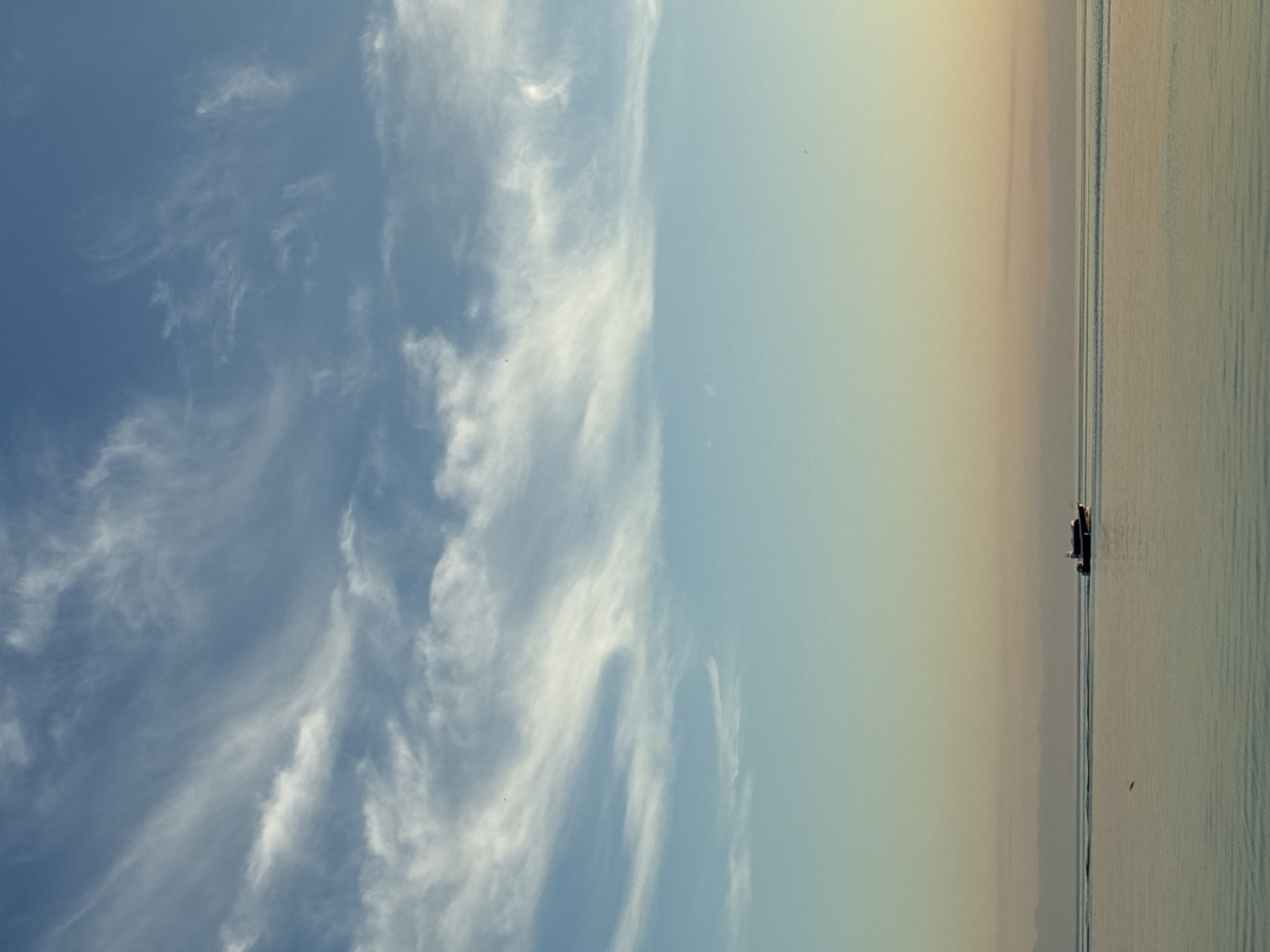An offshore boat travels across a calm body of water under a bright blue sky with wispy white clouds. The boat, a small, dark vessel with a cabin and a person visible at the helm, is positioned on the right side of the image. The water is a muted blue-green, reflecting the sky and distant, hazy landmass visible on the horizon. The transition from the blue sky to the softer, pastel hues of sunset near the horizon creates a serene and tranquil atmosphere. The overall scene is devoid of urban elements, suggesting a natural, open-water environment. While the specific location within Listvyanka, Russia, cannot be definitively determined from the image alone, it depicts a scene typical of the region's extensive waterways, possibly Lake Baikal.