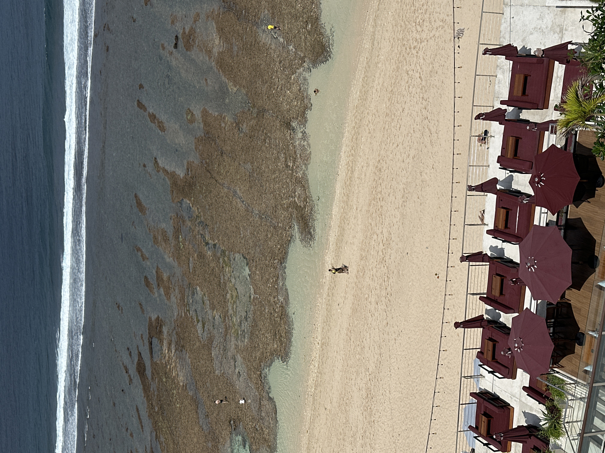 The image shows a beach in Kuta Selatan, Indonesia, with a focus on the shoreline and the area above it. The ocean is visible in the upper portion of the image, with waves breaking on a coral reef. The beach itself is sandy, with a few people walking on it. In the foreground, there is a resort area with lounge chairs and umbrellas. The scene appears to be set during the daytime, with clear weather.